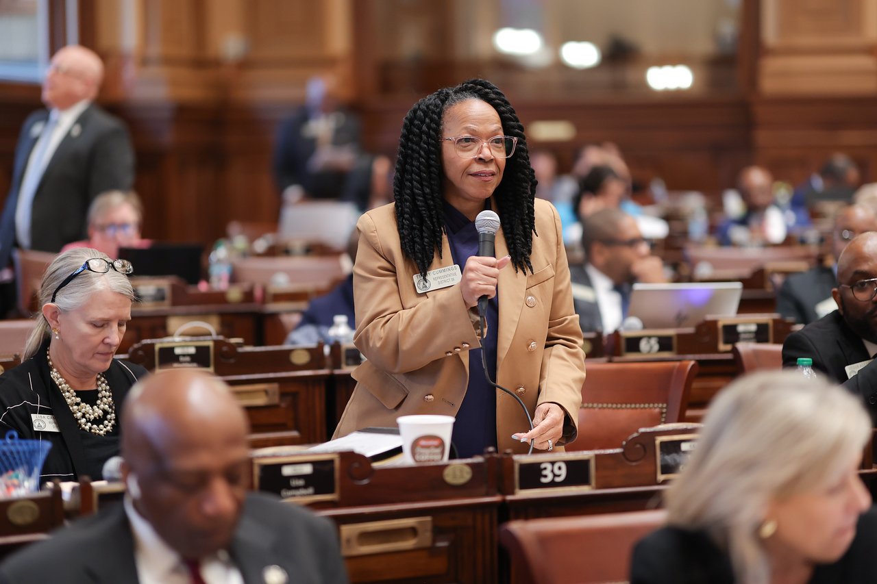 A woman standing and speaking into a microphone in a legislative chamber filled with people, some seated and some working on laptops.