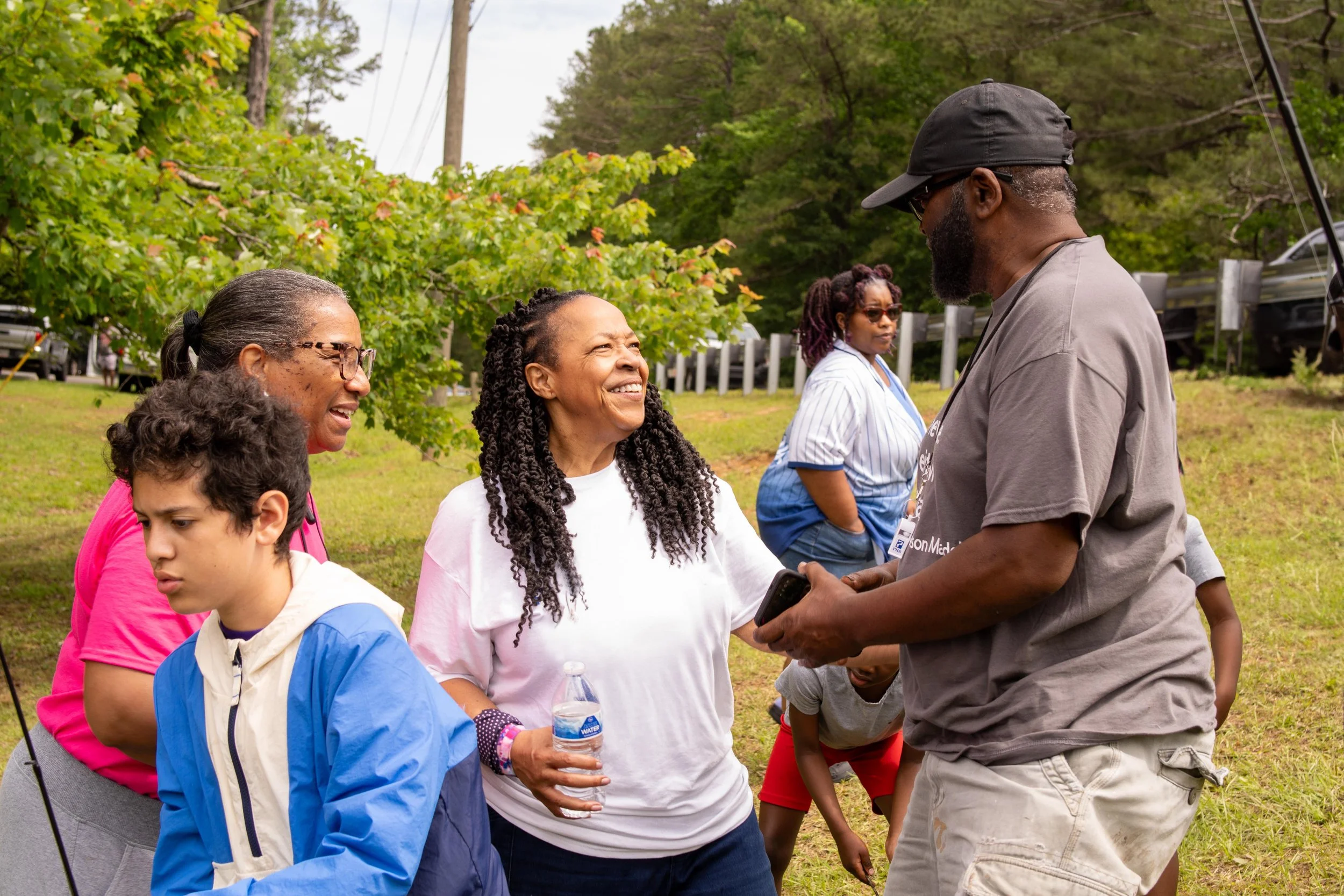 A group of five adults and children standing outdoors. The woman in the center is smiling and holding a water bottle. She is talking to a man wearing a black cap and gray shirt. Other women and children are also present, with green trees and utility poles in the background.