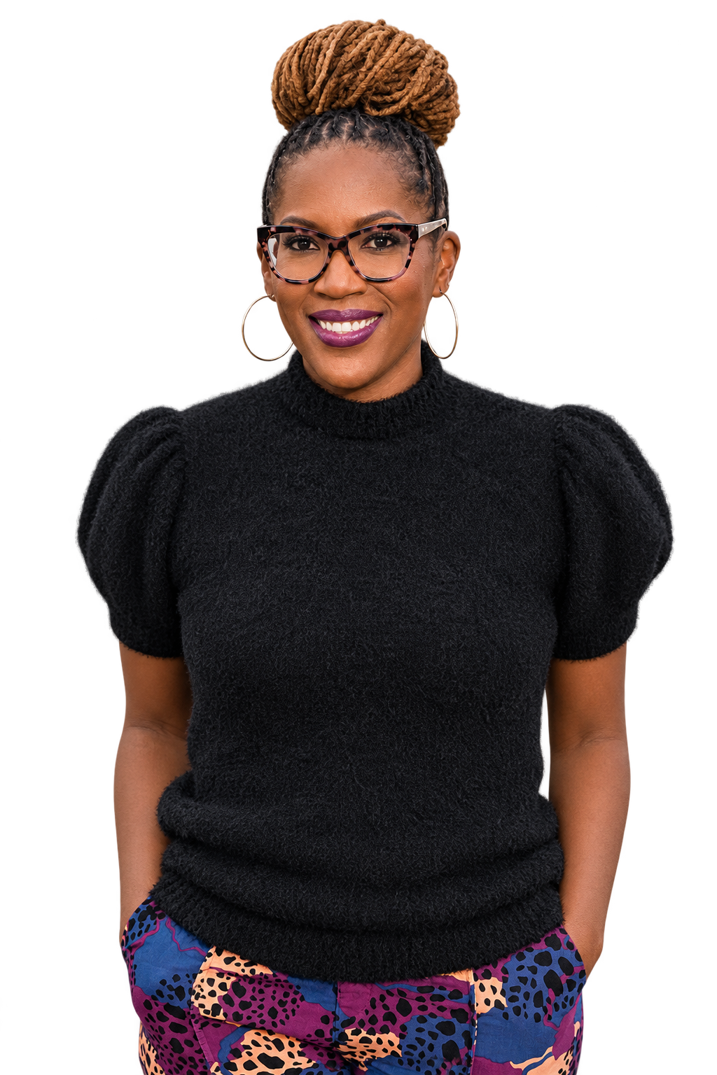 A woman with dreadlocks styled in a bun, wearing glasses, large hoop earrings, a black textured top, and colorful leopard print pants.