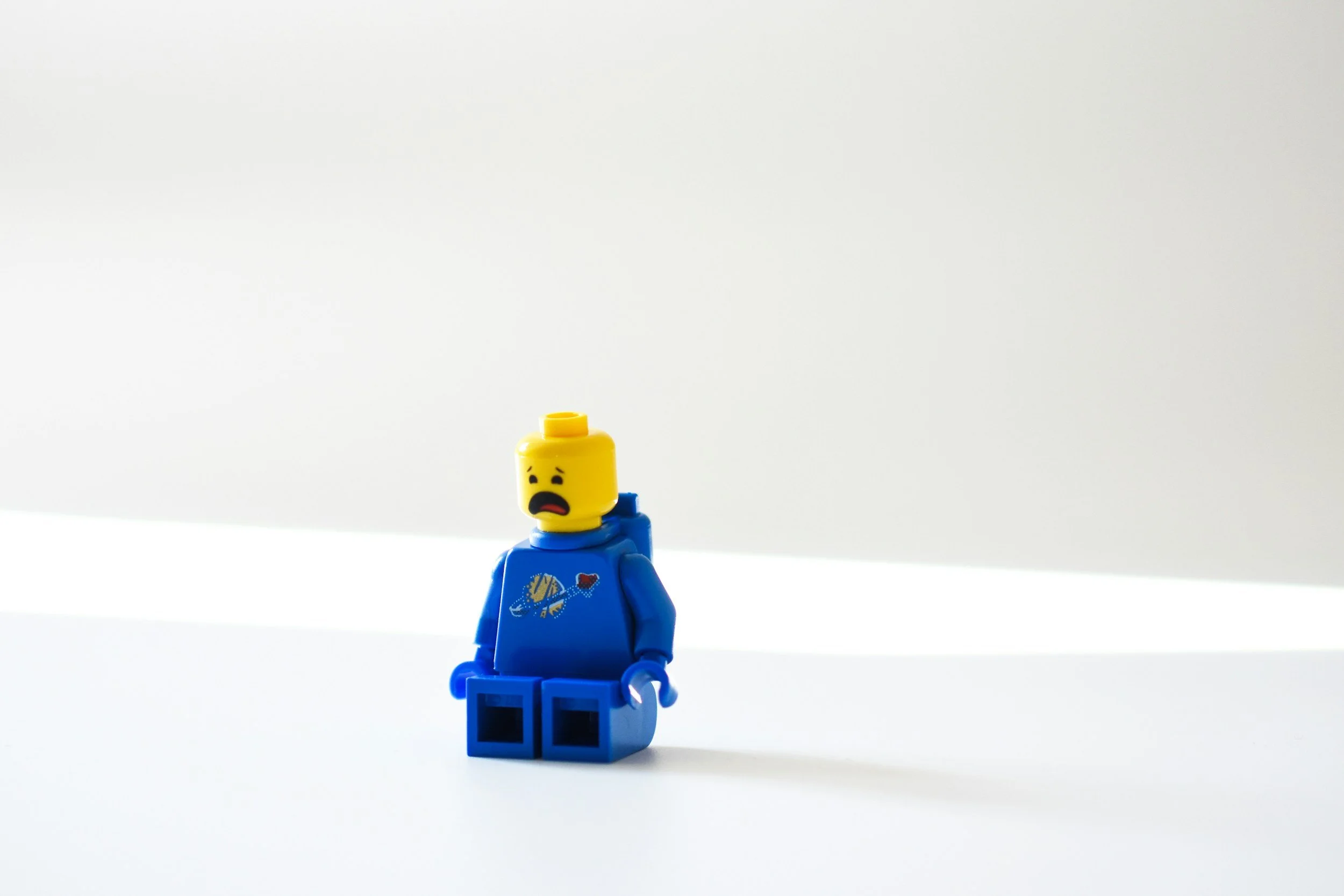 A LEGO minifigure with a yellow head, wearing a blue hoodie with a space-themed design, standing alone on a white surface against a plain white background. The figure's facial expression looks distressed or worried.