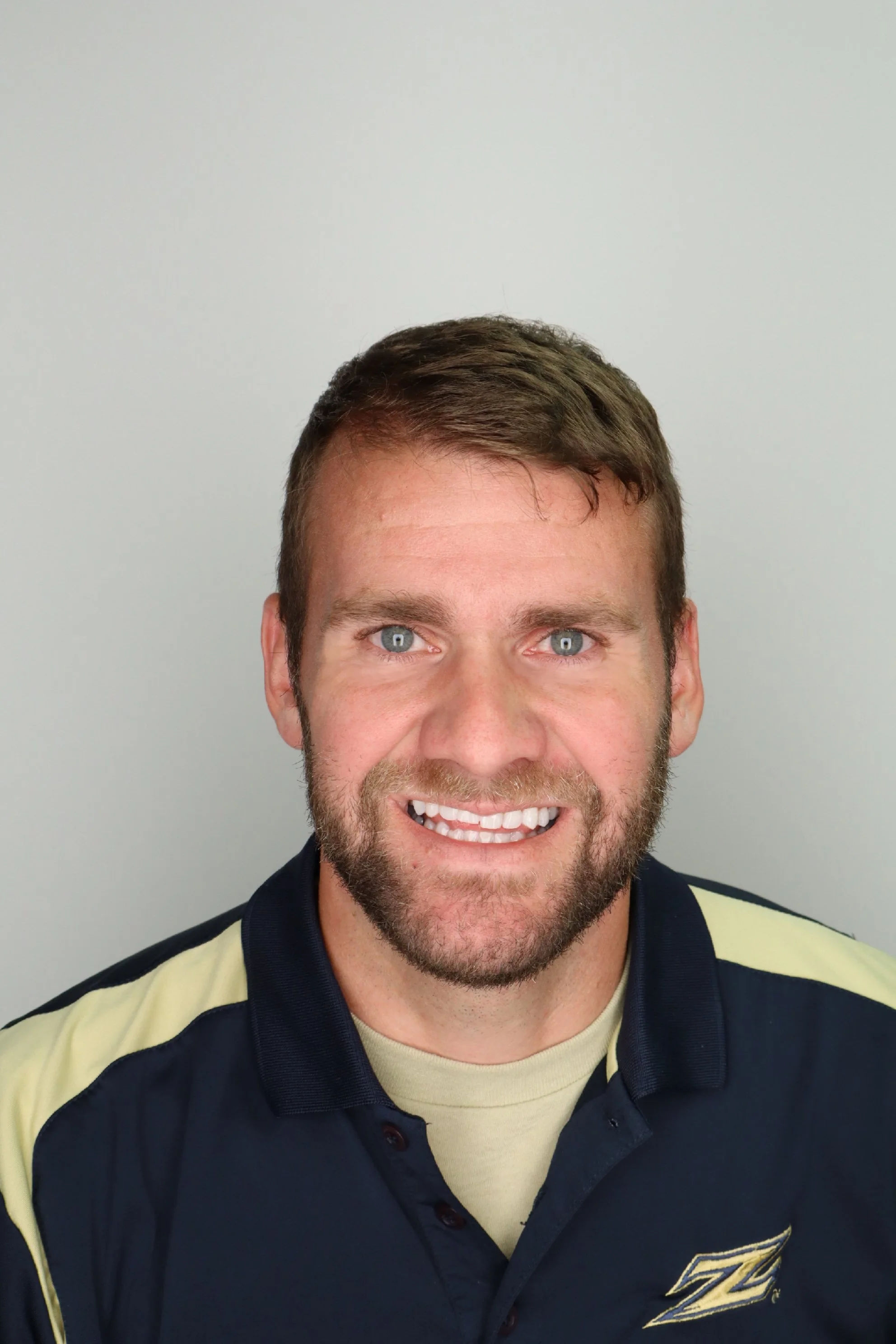 A smiling man with short brown hair, beard, and blue eyes wearing a dark navy and yellow sports shirt standing against a plain light gray background.