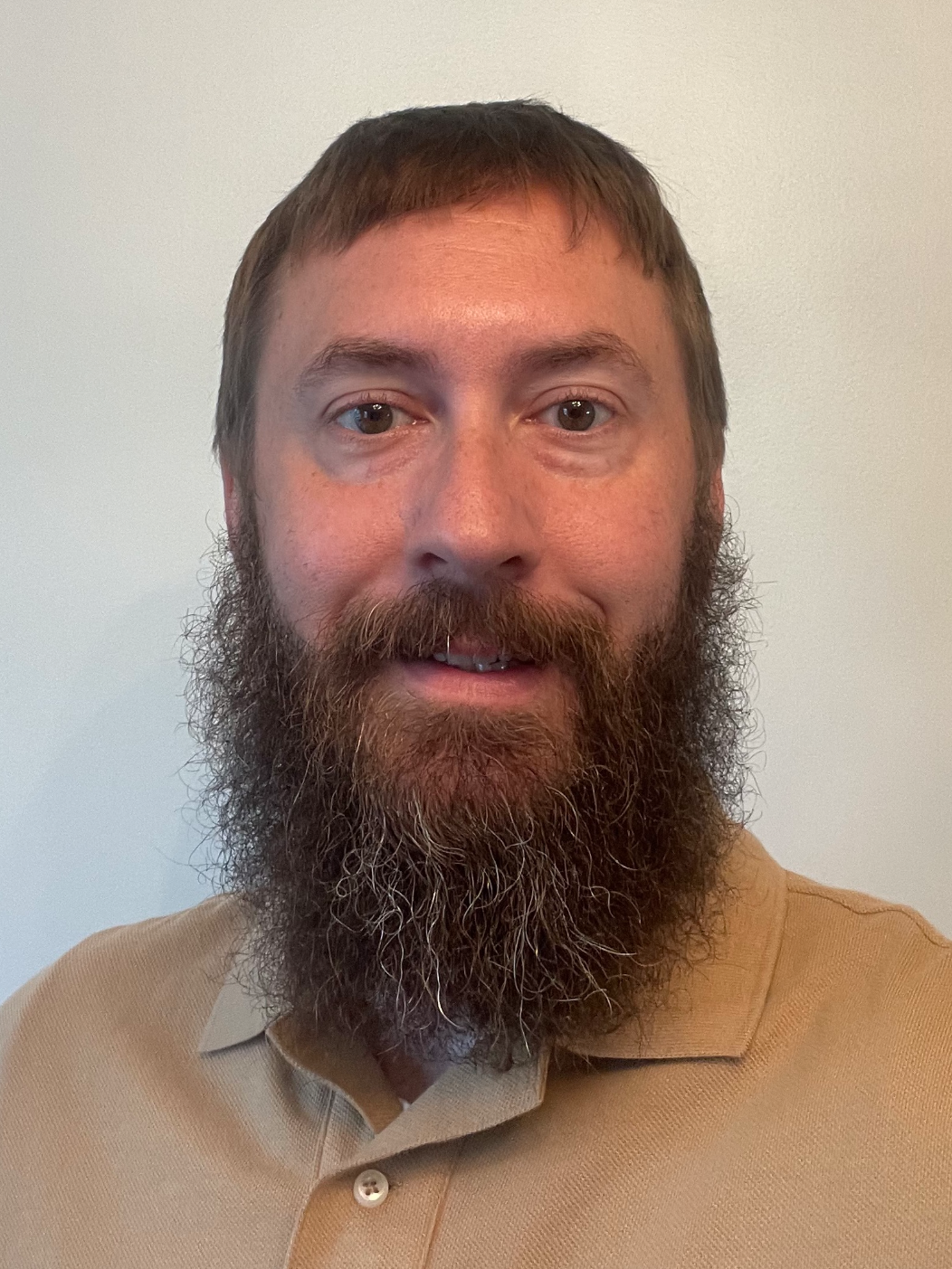 A man with brown hair, a full beard, and brown eyes, wearing a beige polo shirt, smiling and looking directly at the camera against a plain white background.