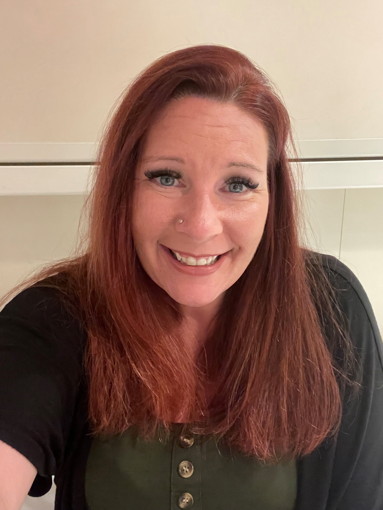 A smiling woman with long red hair, blue eyes, and a nose piercing, wearing a black shirt with buttons, taking a selfie in front of a beige background.