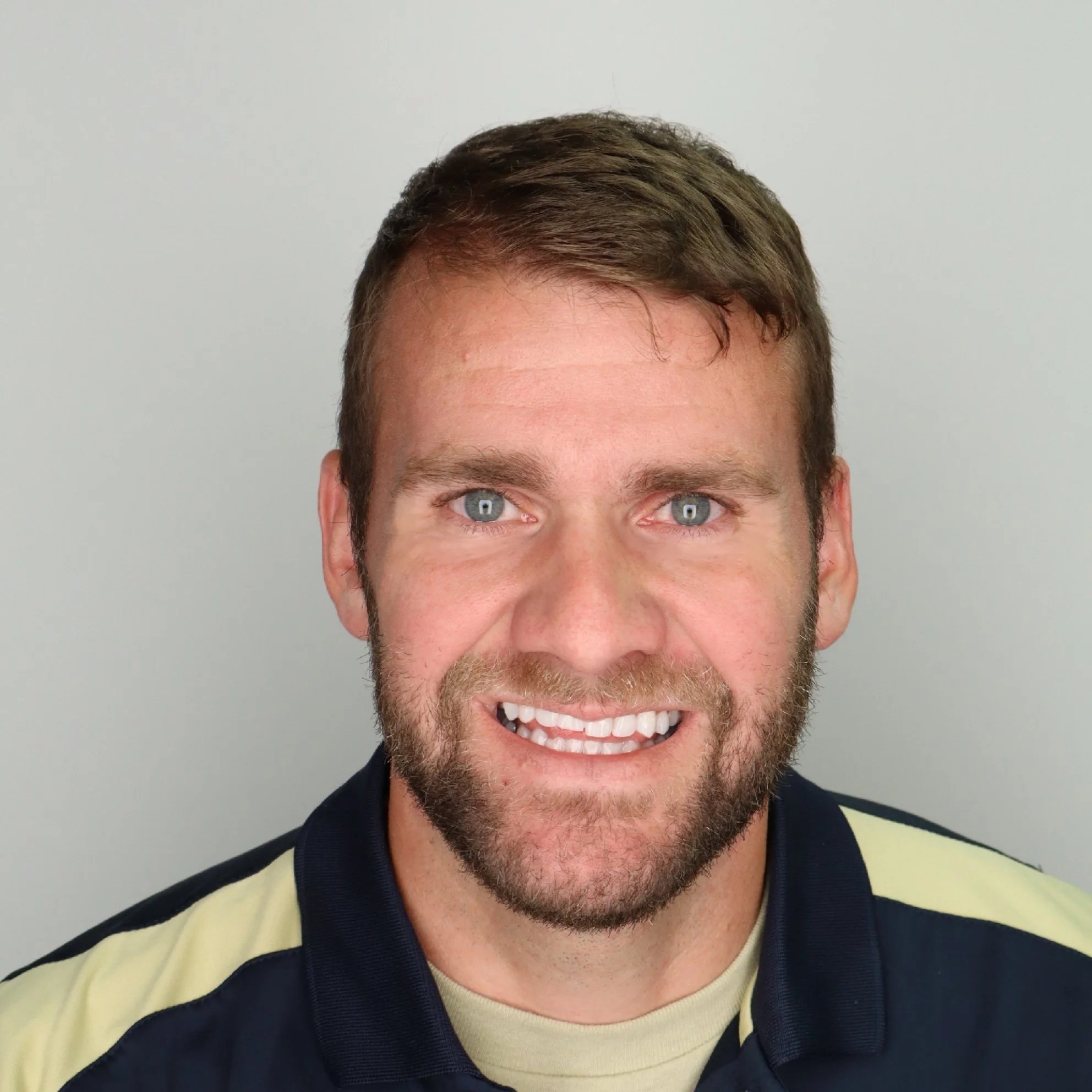 Close-up of a smiling man with blue eyes, brown hair, and light stubble, wearing a black and yellow sports jersey.