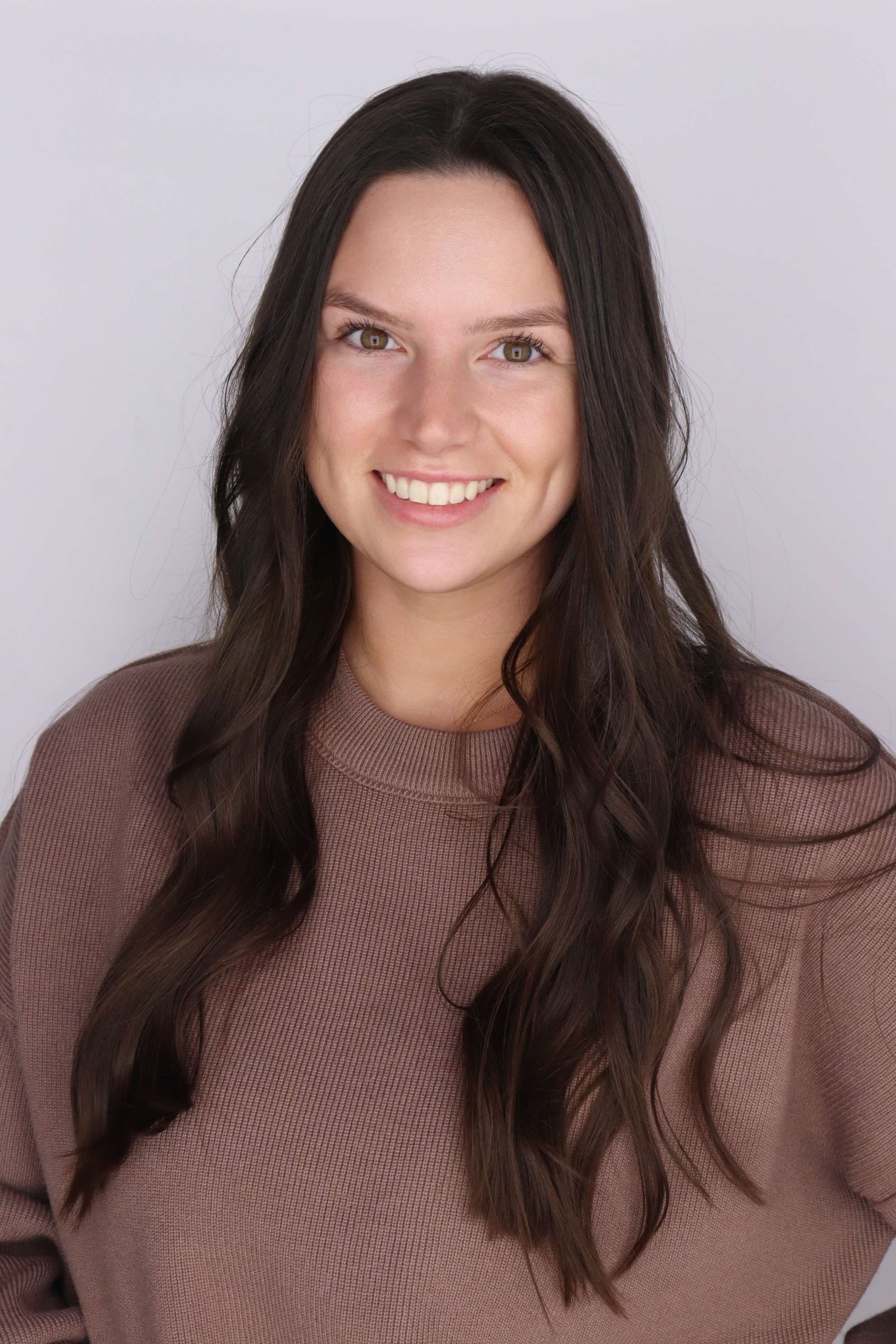 Close-up of a young woman with long wavy brown hair, wearing a brown long-sleeve shirt, smiling against a plain white background.