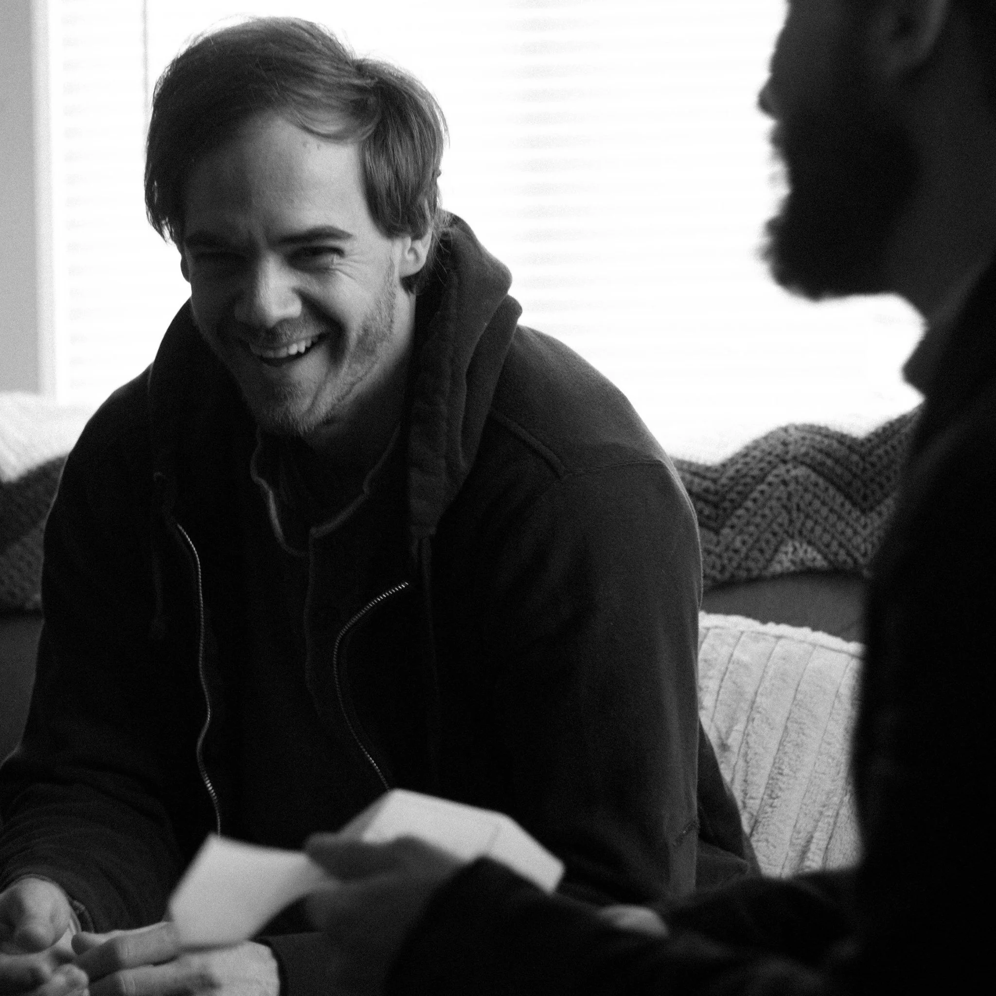 A black and white photo of two smiling people facing each other, one with short wavy hair and the other with darker skin and curly hair, sitting close indoors.