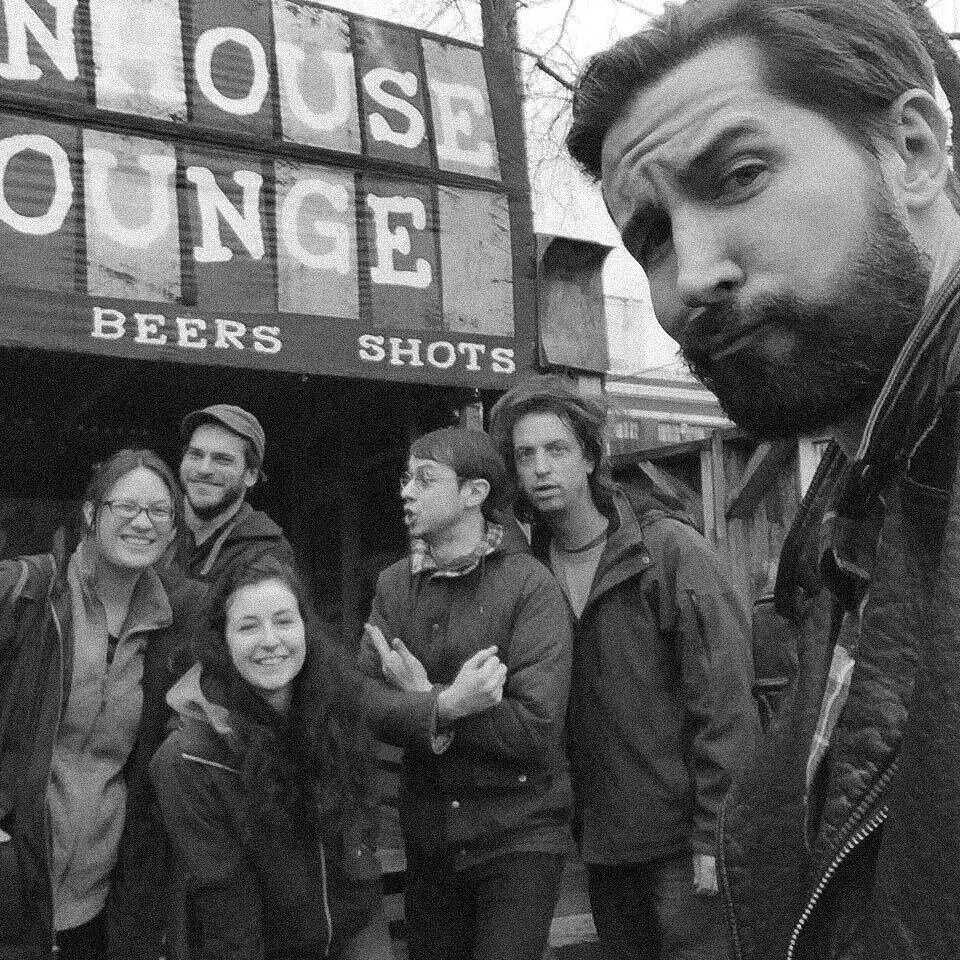 A group of five young adults posing outside a building with a sign that reads 'NOUSE BINGE BEERS SHOTS'. One person is taking a selfie, and the others are smiling or making expressive faces.
