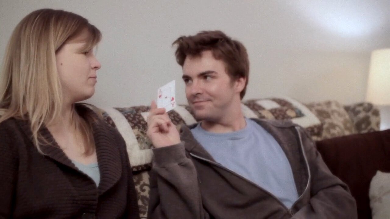 A young man holding a playing card with two hearts while sitting on a sofa next to a young woman, in a living room.