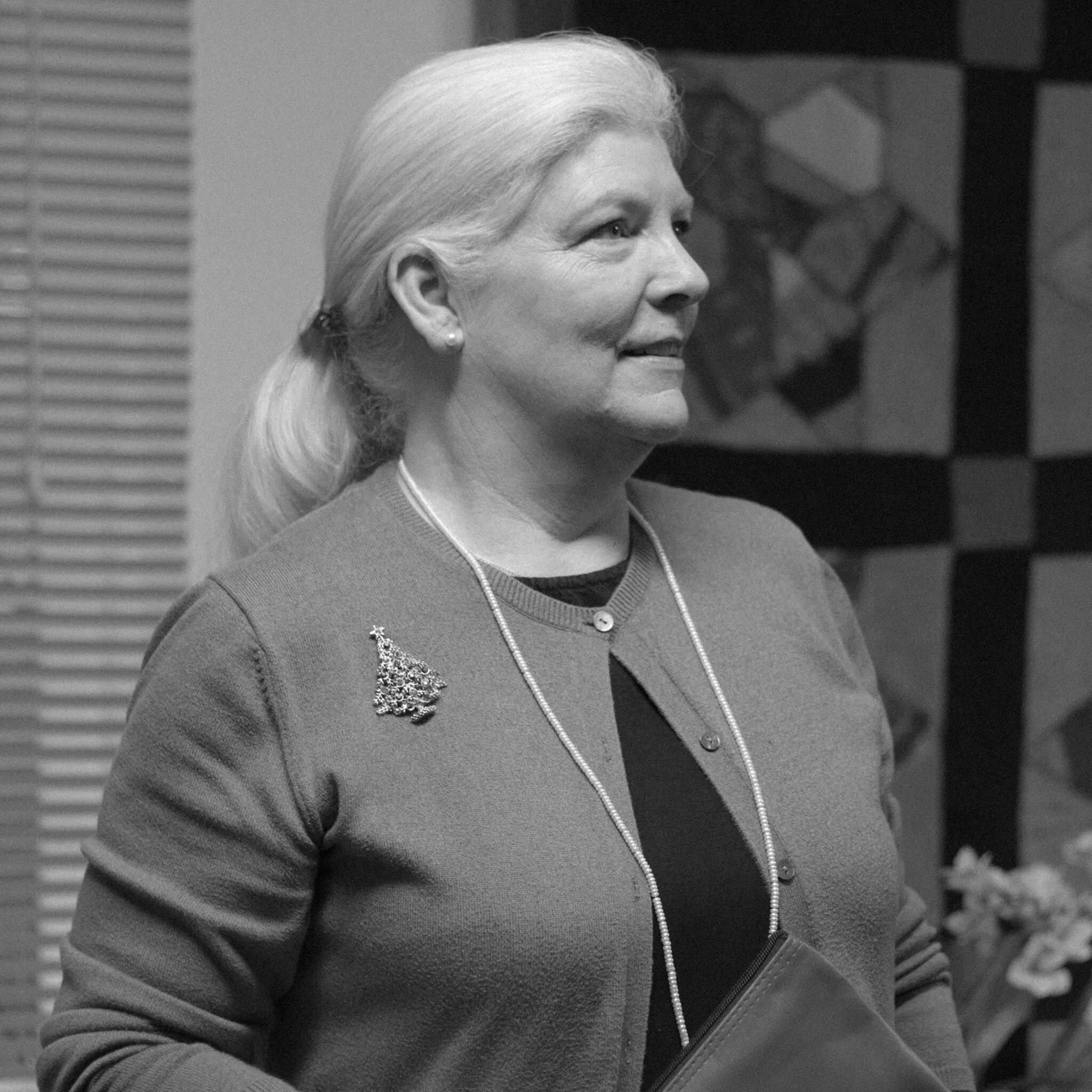 Side profile of a smiling woman with blonde hair tied back, wearing pearl earrings, a necklace, and a blazer with a brooch, indoors with modern decor.