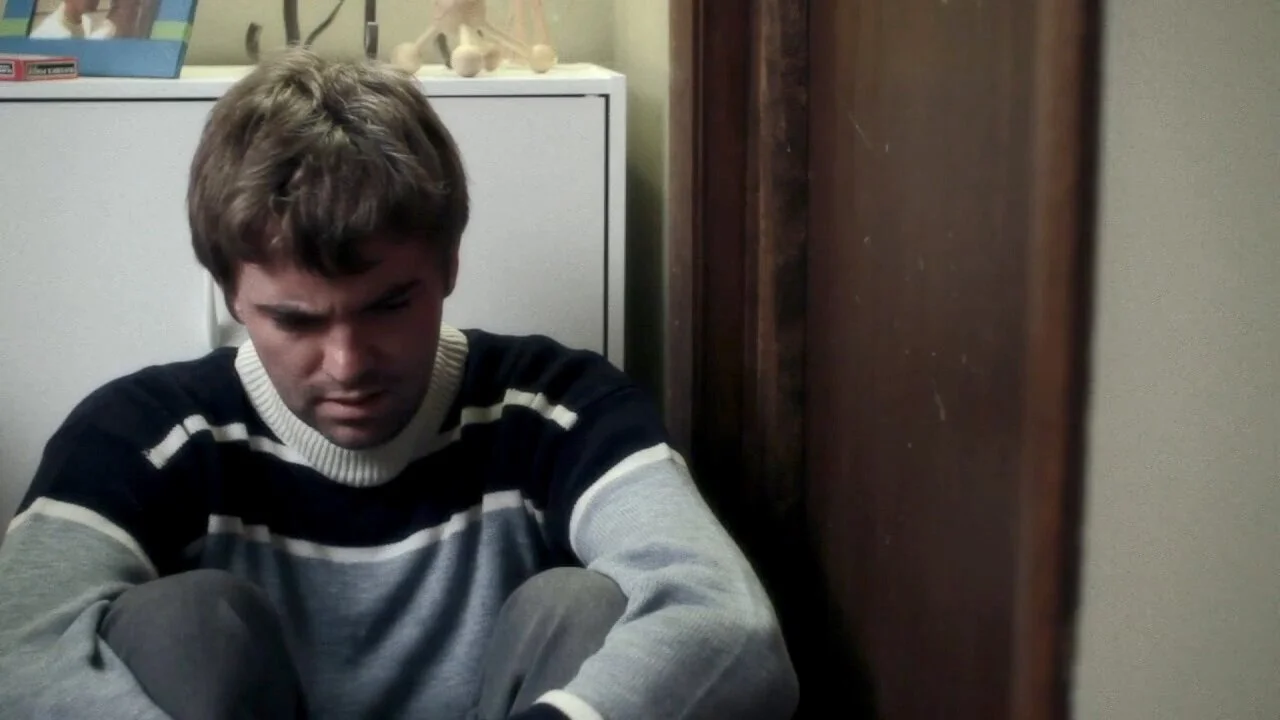 Young man sitting on the floor with a pained or distressed expression in a room with a white cabinet and various objects in the background.