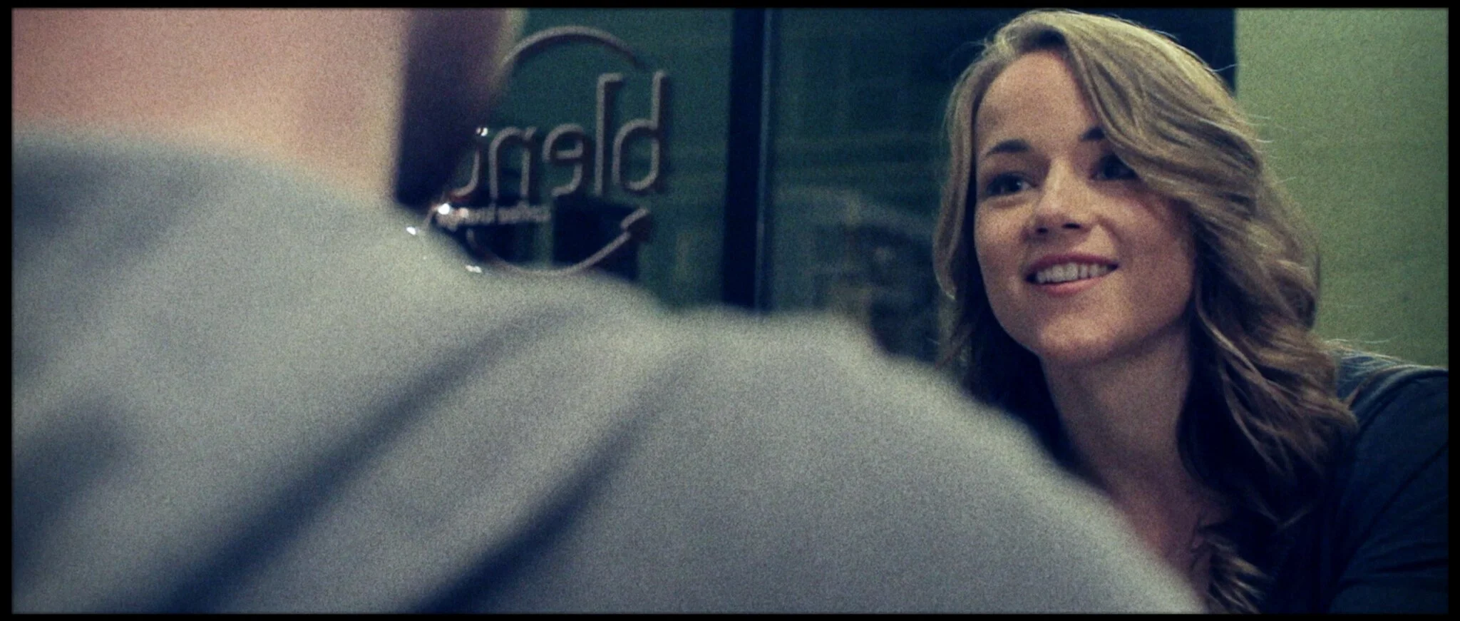 A woman with long, wavy hair smiling and looking at a person in the foreground, with a blurred gray and green background.