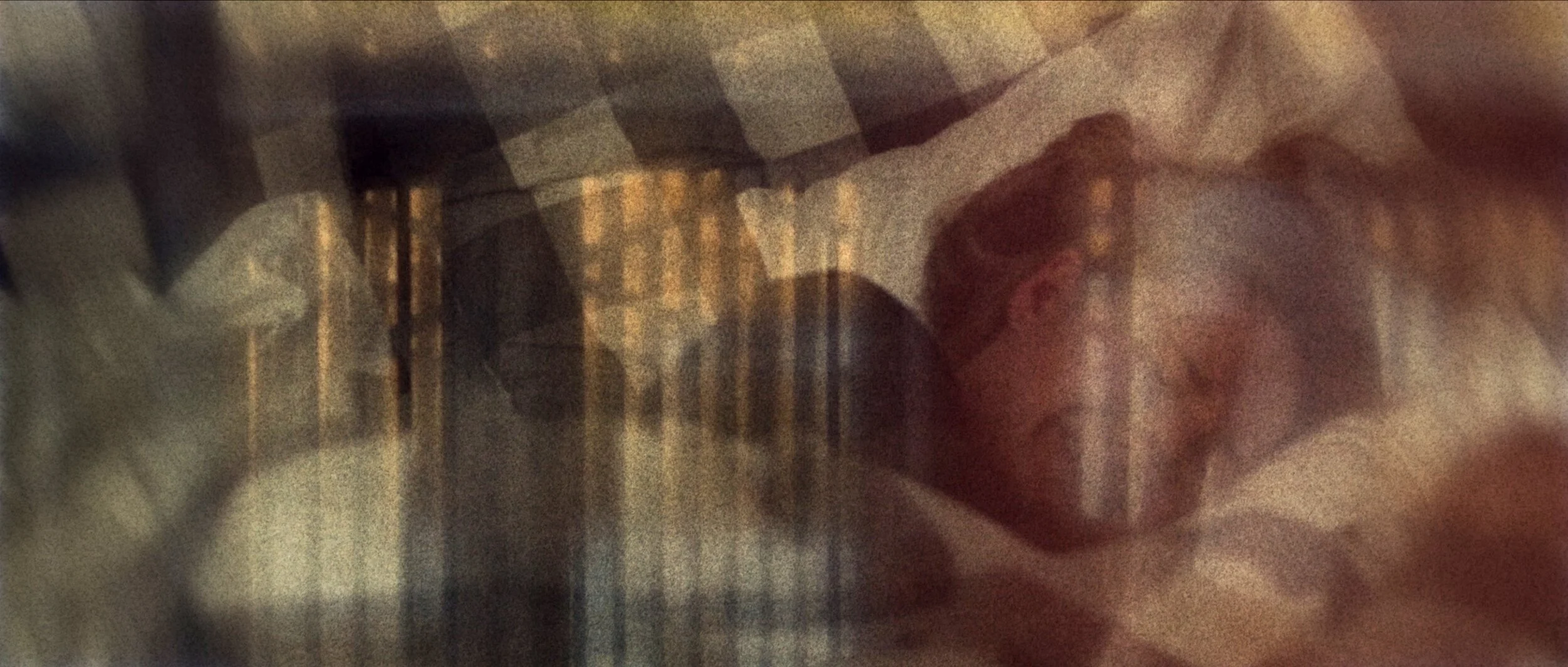 A person wearing a plaid shirt appears to be lying down, possibly covered with a blanket or sheet. The image is blurry and difficult to see clearly.