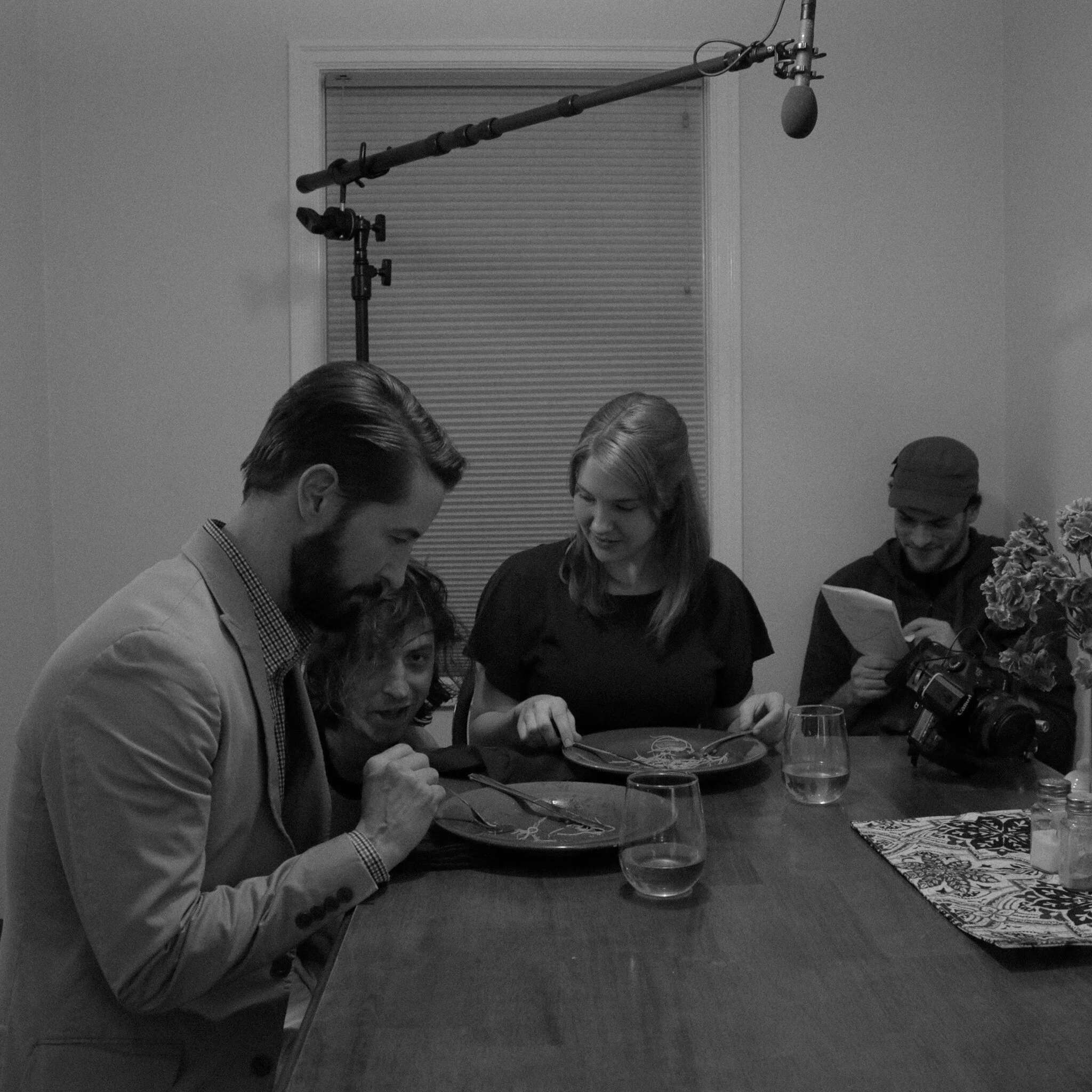 Black and white photo of people sitting around a table at a gathering or dinner event. Some are engaged in conversation, and a woman is holding a flower bouquet. There are glasses and plates on the table, and a microphone is suspended above the scene