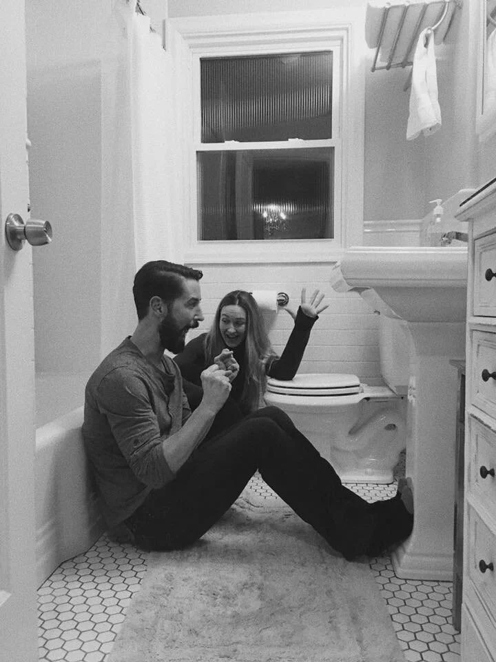 Two people sitting on the bathroom floor, smiling and interacting, with a toilet, sink, and window in the background.