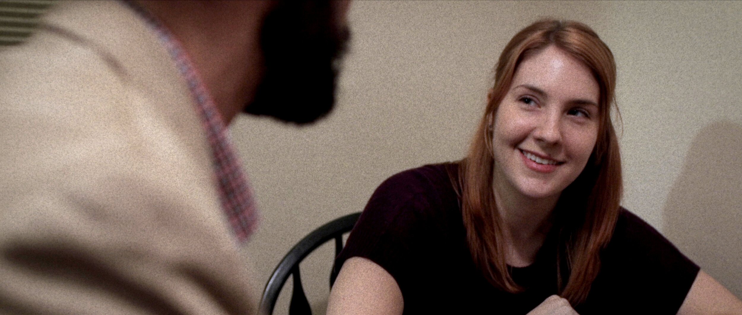 A woman with red hair smiling while talking to a man in a beige jacket and wearing a checkered shirt. They are sitting at a table with a plain wall in the background.