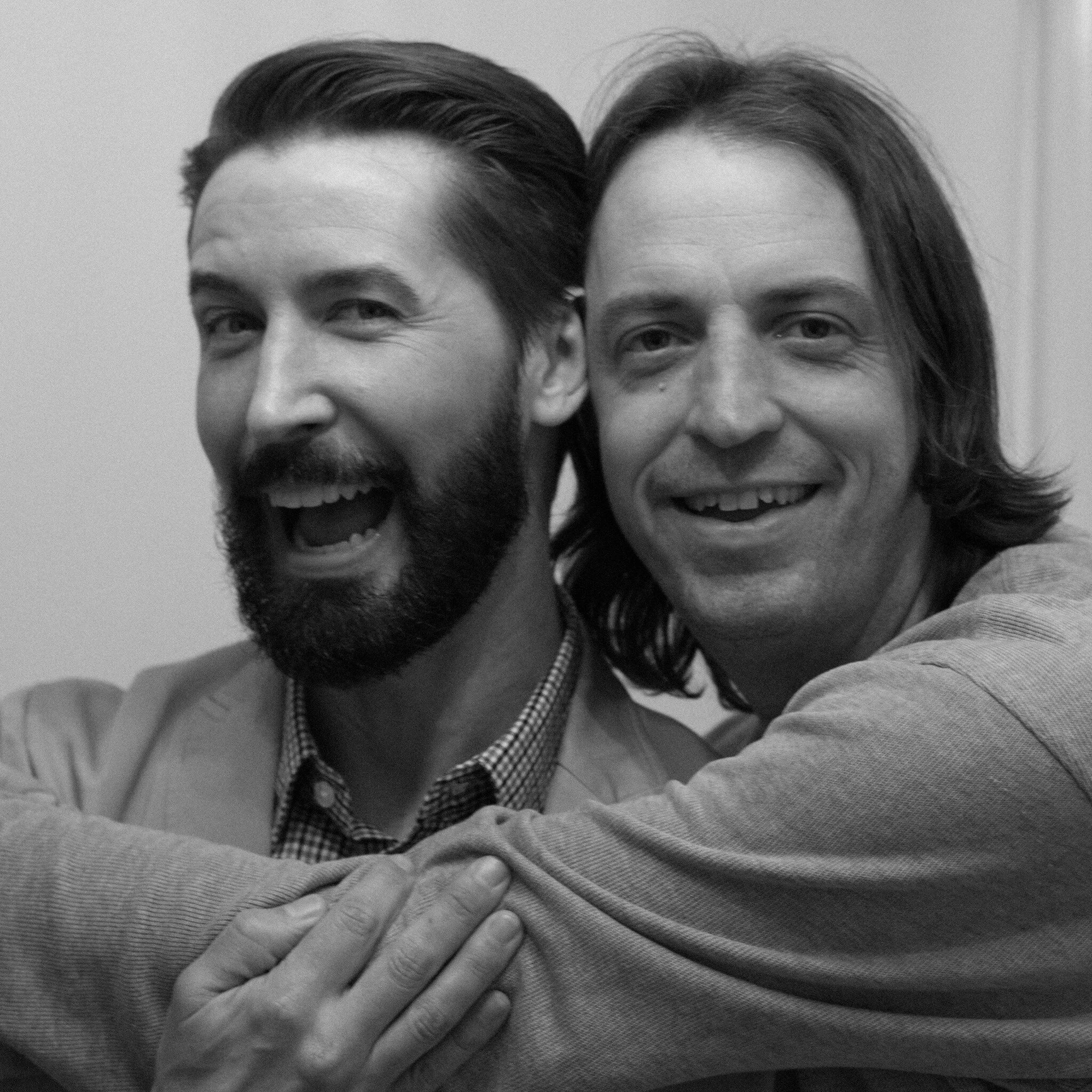 Close-up of two people smiling, one with a beard and the other with long hair, in black-and-white.