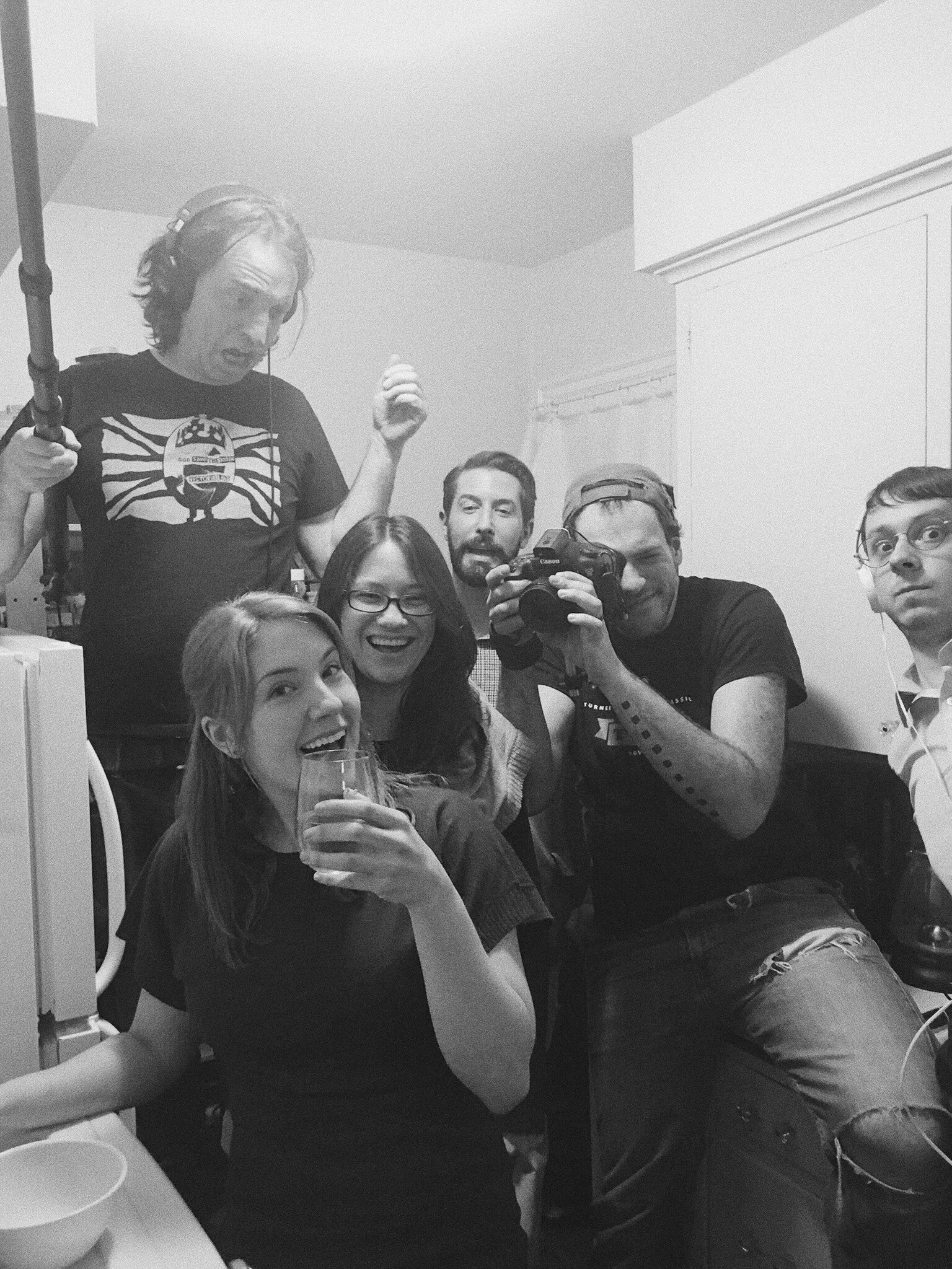 A group of people gathered in a room, with one woman holding a glass and smiling at the camera, while several others pose and take a picture. The environment appears to be informal and social.