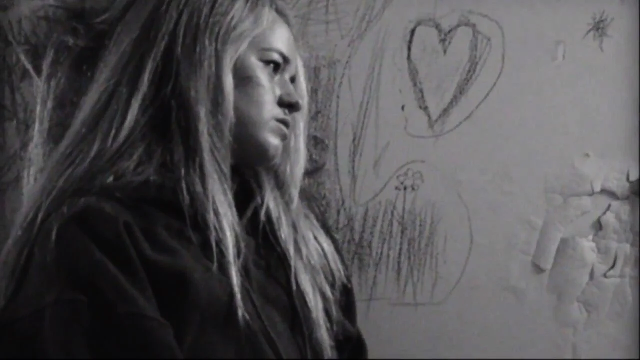 A woman with long, messy hair looking to the side, standing next to a wall with a heart and some other drawings, in black and white.