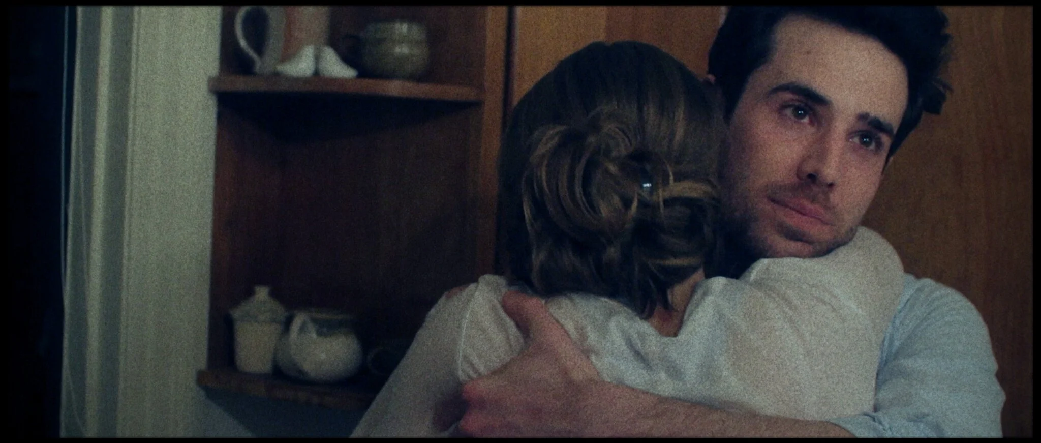 A young man and a young woman hugging each other warmly in a cozy indoor setting. The man has dark curly hair and is gazing softly, while the woman has wavy hair and is hugging him tightly. Background includes wooden shelves with decorative items.