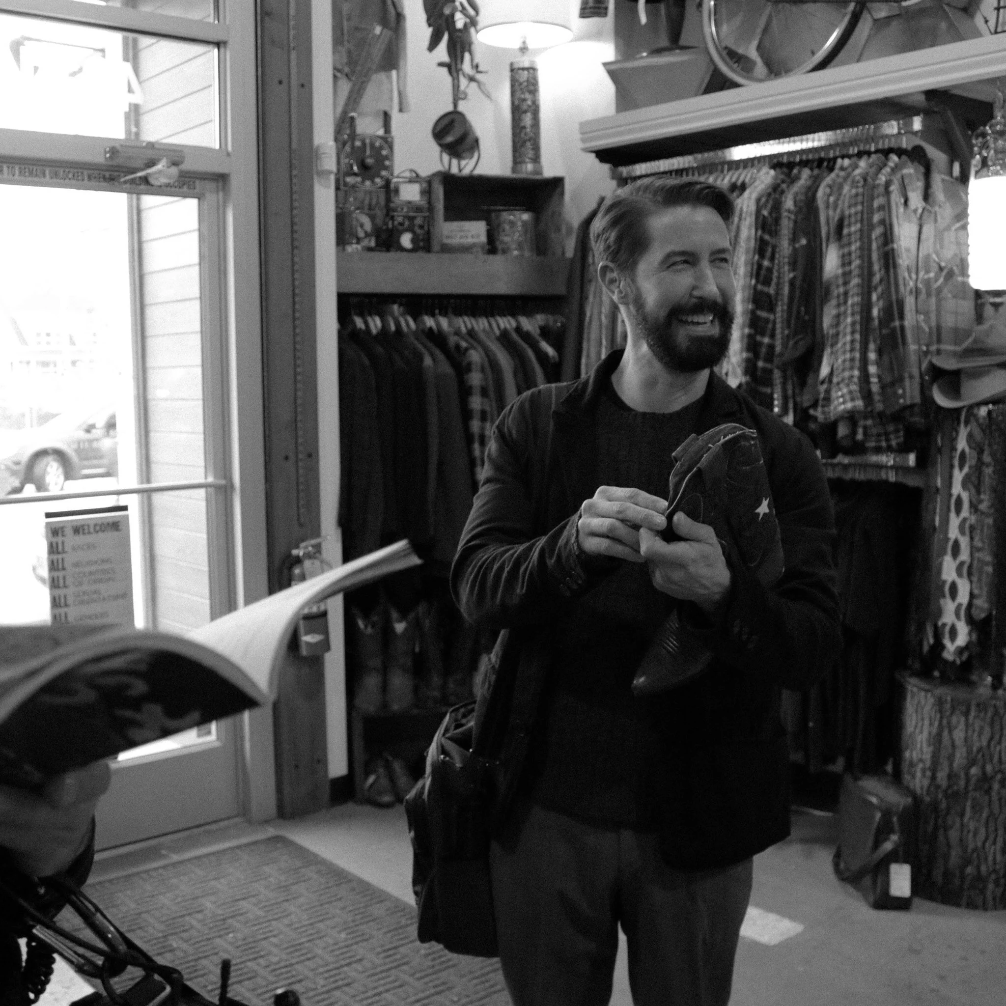 A smiling man with a beard and styled hair, wearing a dark jacket, is looking to the right inside a clothing store. Behind him are hanging clothes, shelves with various items, a window with partially open blinds, and a stroller partially visible in t