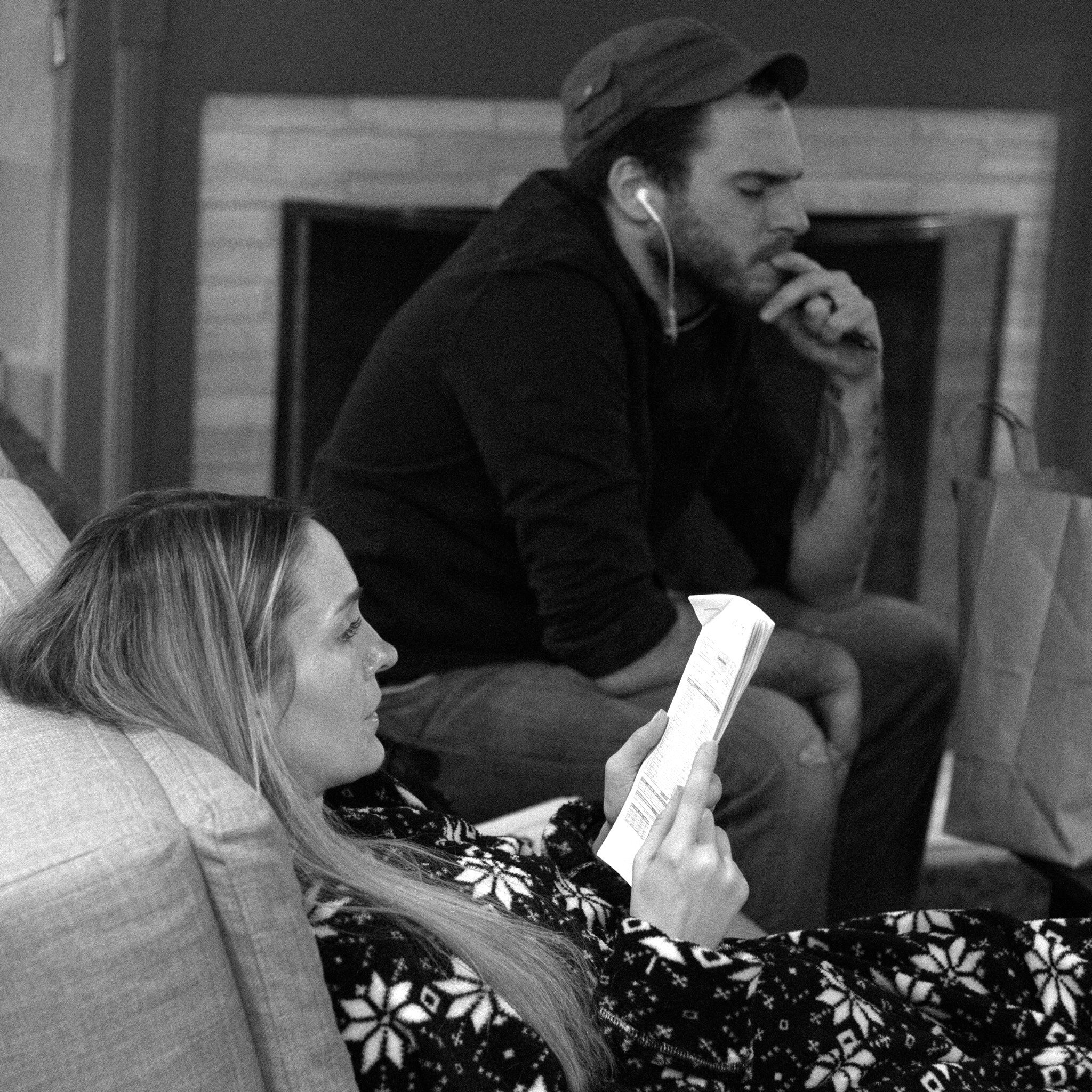 A person sitting on a sofa, reading a booklet, with blonde hair and wearing a dark patterned dress. Another person is partially visible in the foreground, with only their hair showing.