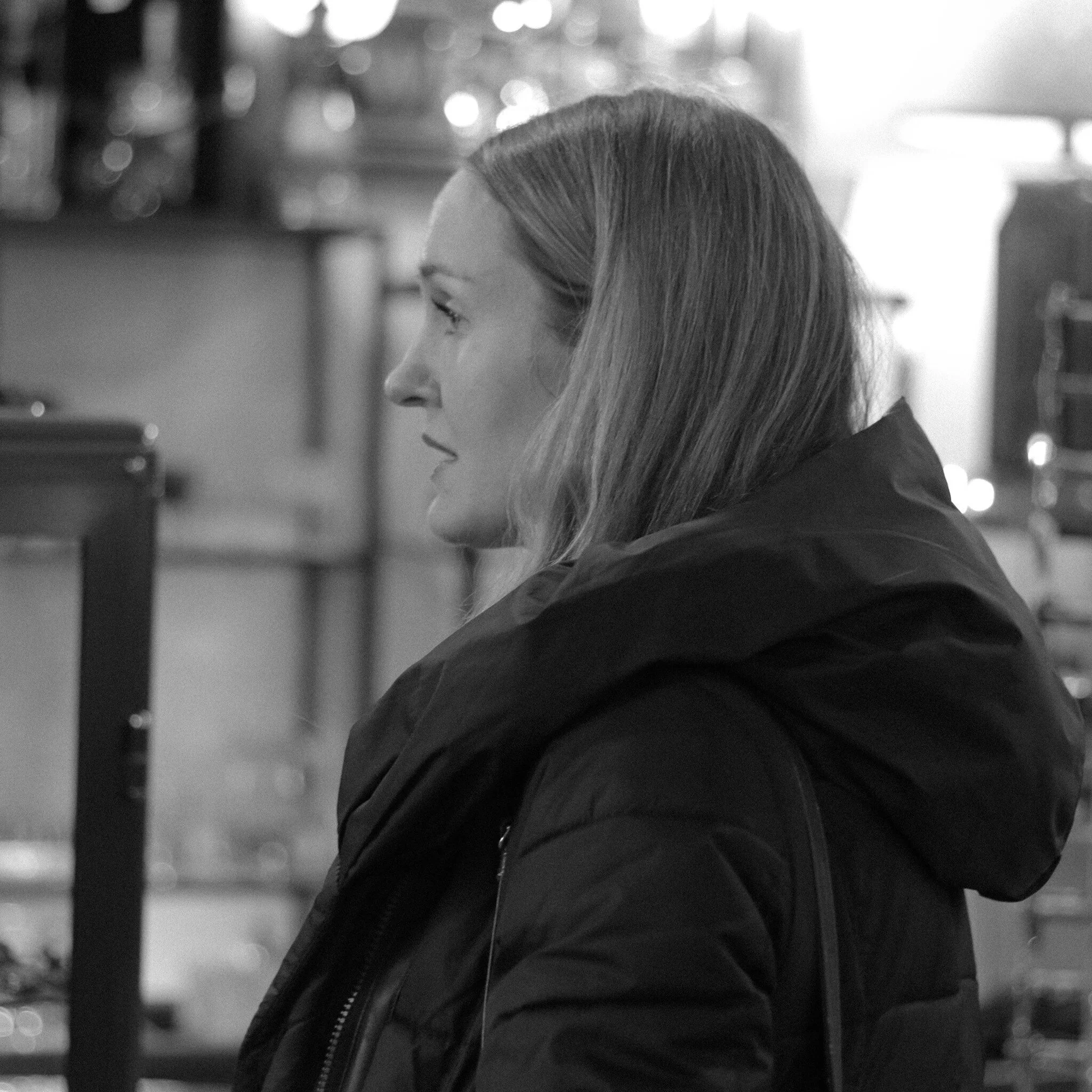 Black and white photo of a woman with long hair, wearing a dark jacket, and looking to the side, in front of a background with blurry lights.