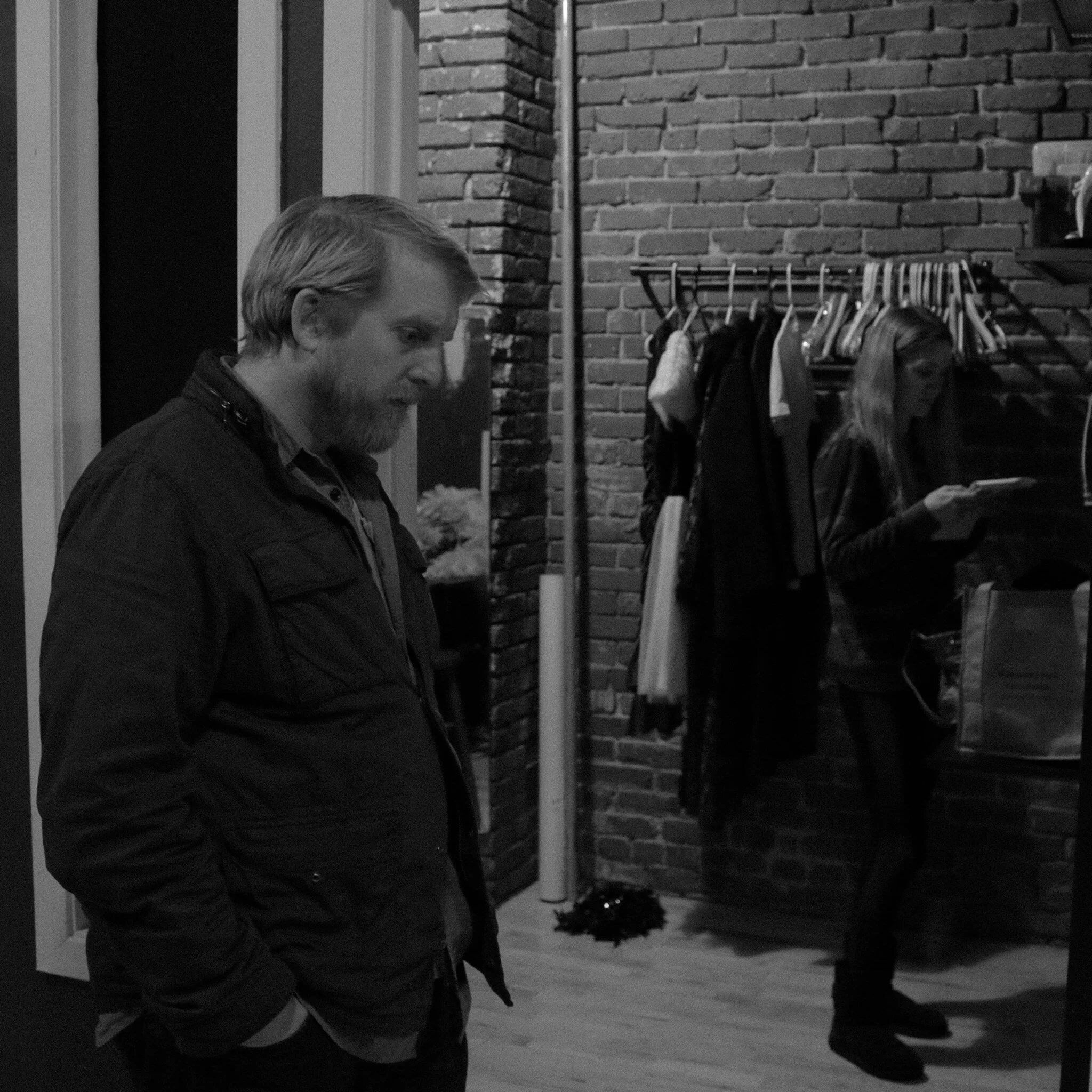 A black and white photo of a man with a beard and short hair standing indoors with brick walls behind him. There is clothing hanging on a rack and a small Christmas tree on the floor.