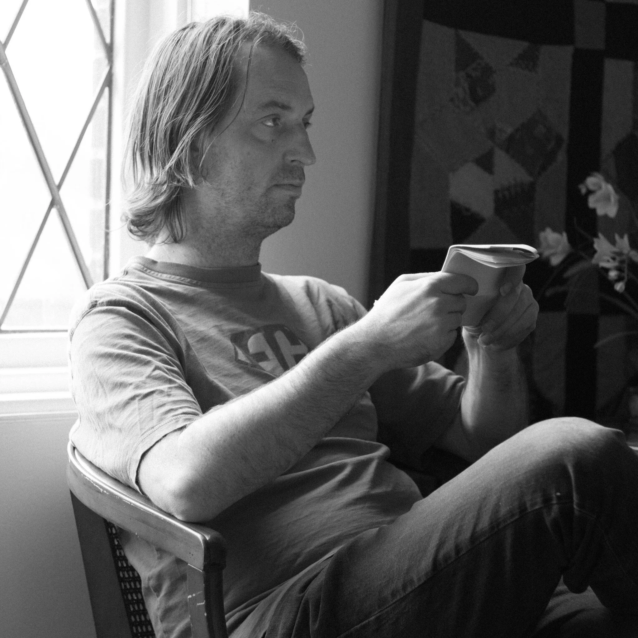 A black and white photo of a man with shoulder-length hair, sitting indoors and holding a book or tablet.