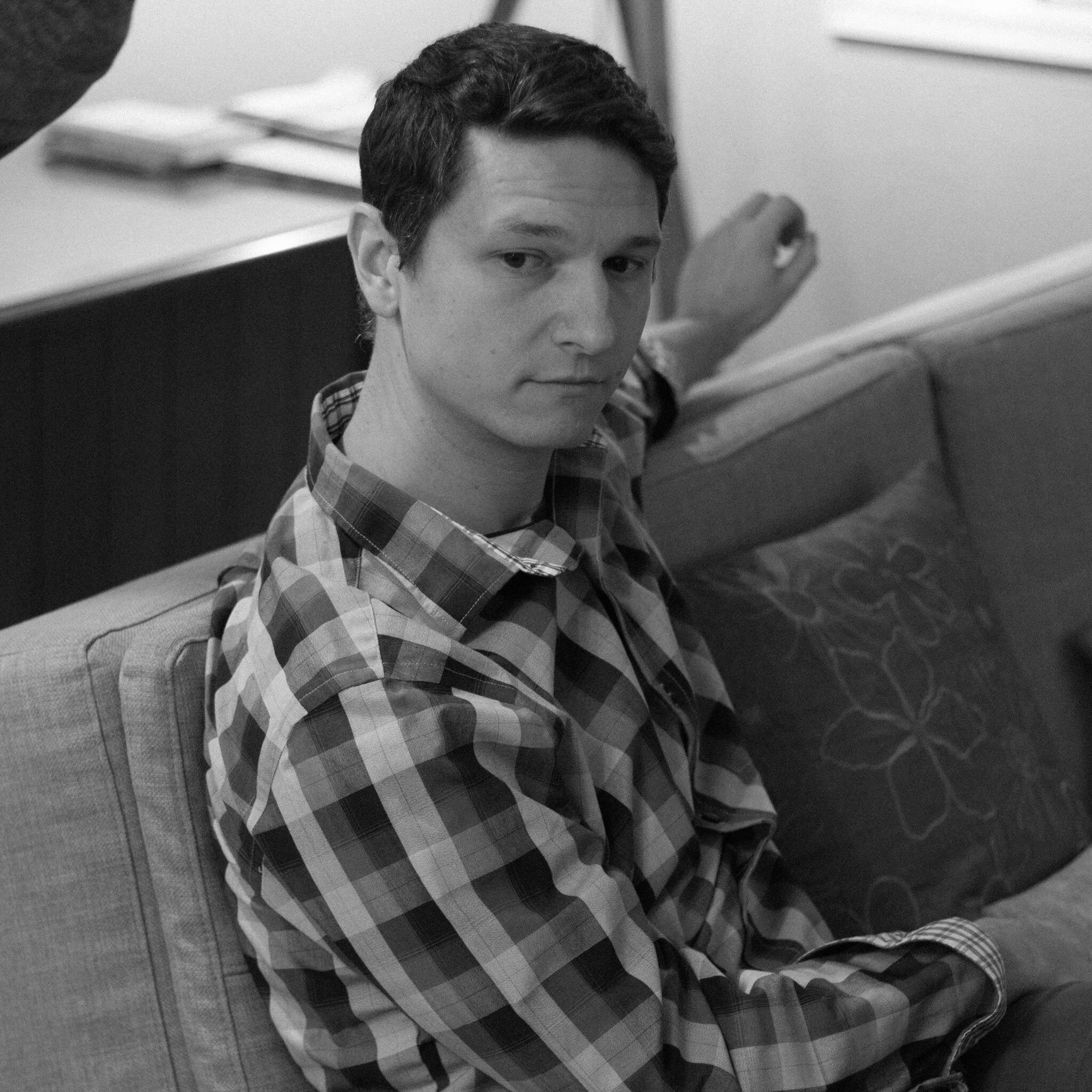 A young man with dark hair and a checkered shirt looking directly at the camera while sitting on the floor next to a person with a rolled-up document or poster.