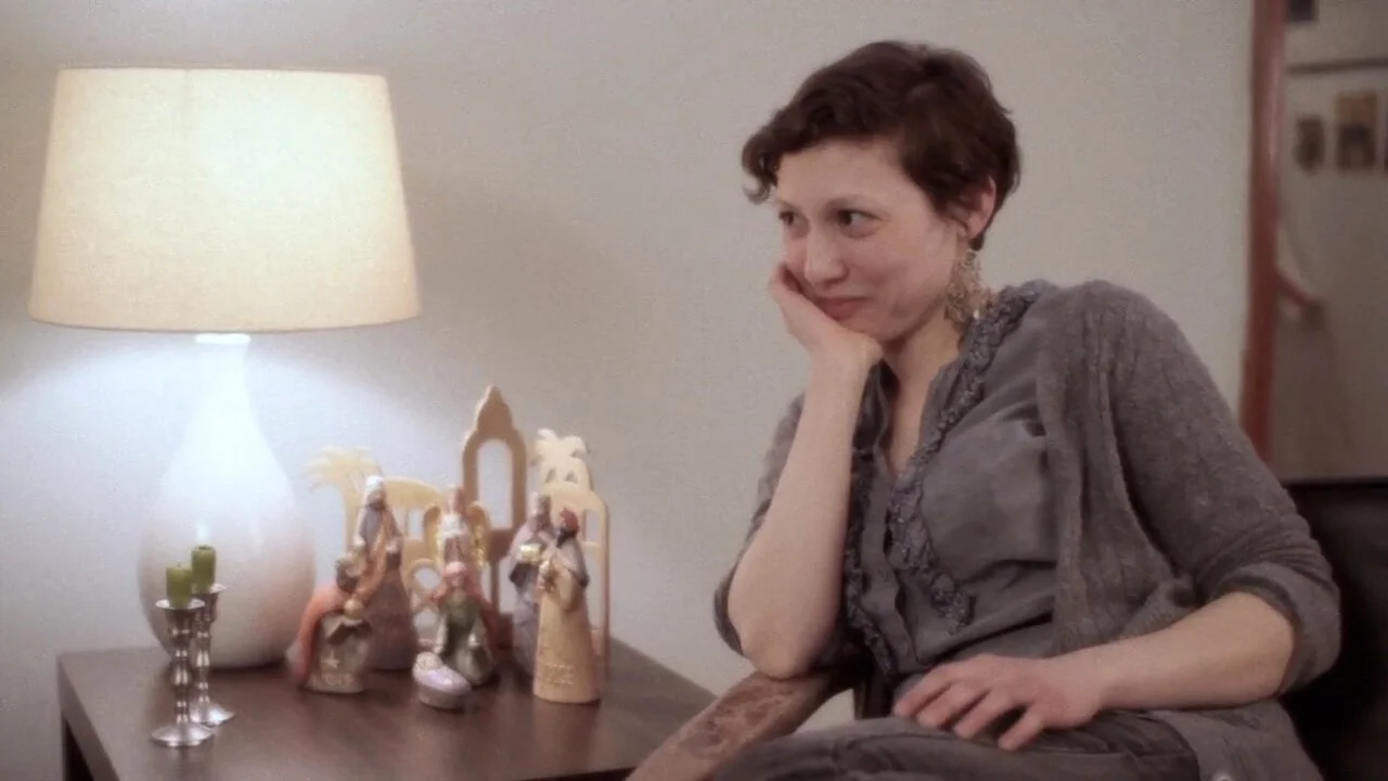A woman with short curly hair sitting at a table with her chin resting on her hand, looking to her right, with a lit lamp and nativity scene figurines on the table beside her.