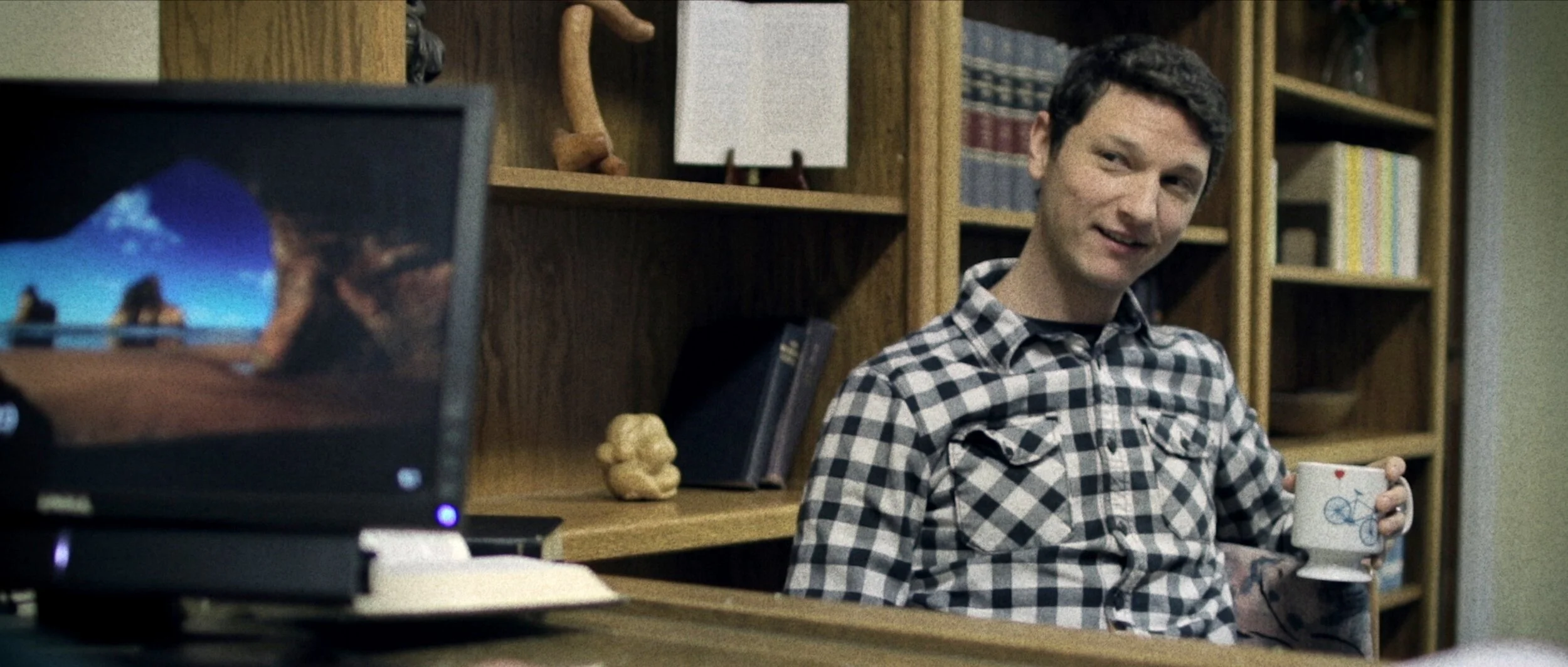 A young man with short dark hair, wearing a black and white plaid shirt, is sitting at a desk holding a white mug with a bicycle drawing. There are bookshelves with books and decorative objects behind him, and a computer monitor is in the foreground.