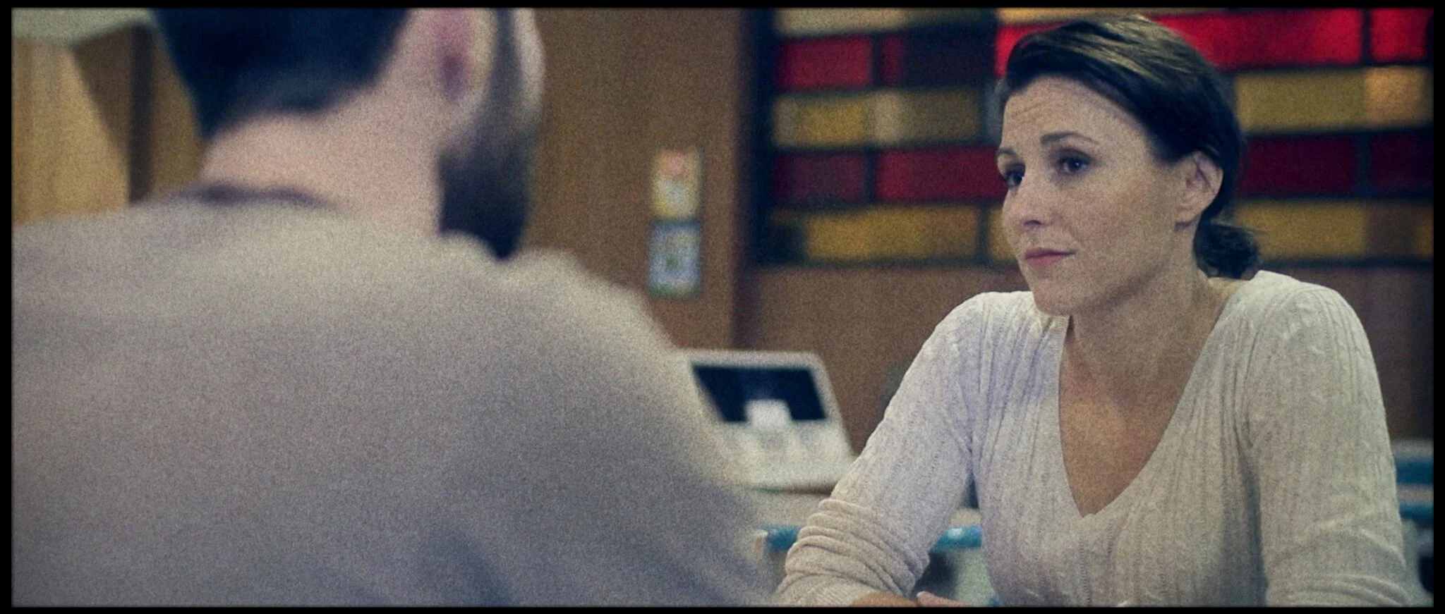 A woman with short brown hair wearing a white sweater sits at a table and looks at a man across from her, who is blurred and facing away from the camera. The background has a colorful wall.