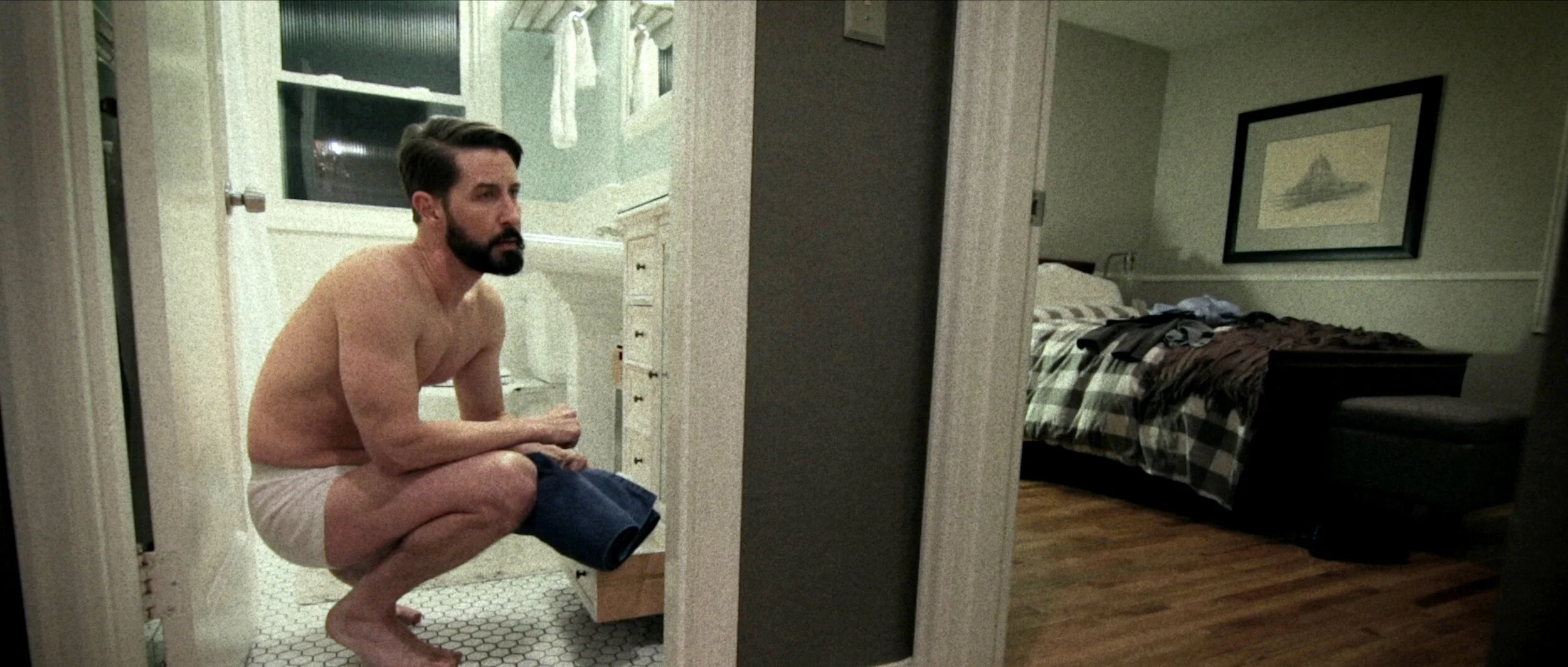 A man with a beard, shirtless, crouching in a bathroom, holding a pair of jeans, and looking to his left. The bathroom is next to a bedroom with a bed and a framed picture on the wall.