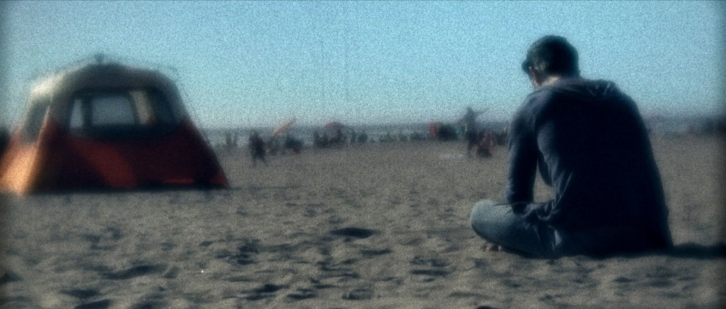 A person sits on the sand at the beach, facing away from the camera, near a tent with other beachgoers in the background.