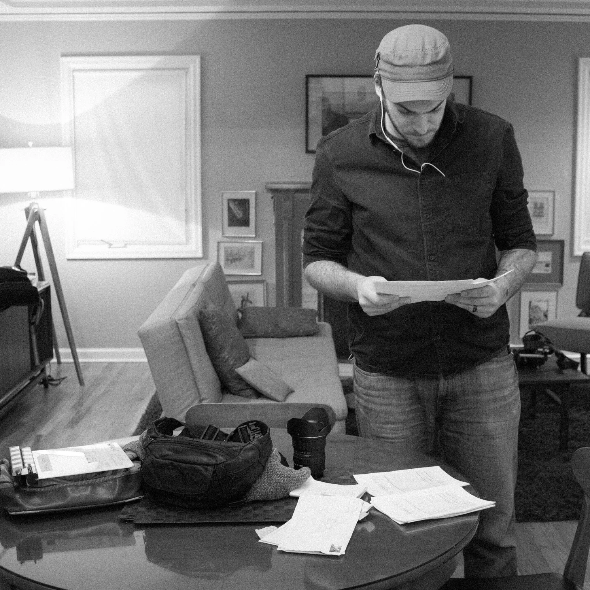 A person wearing a cap and earphones, looking down at papers on a desk, with a backpack and books nearby, in a room with artworks and a window.