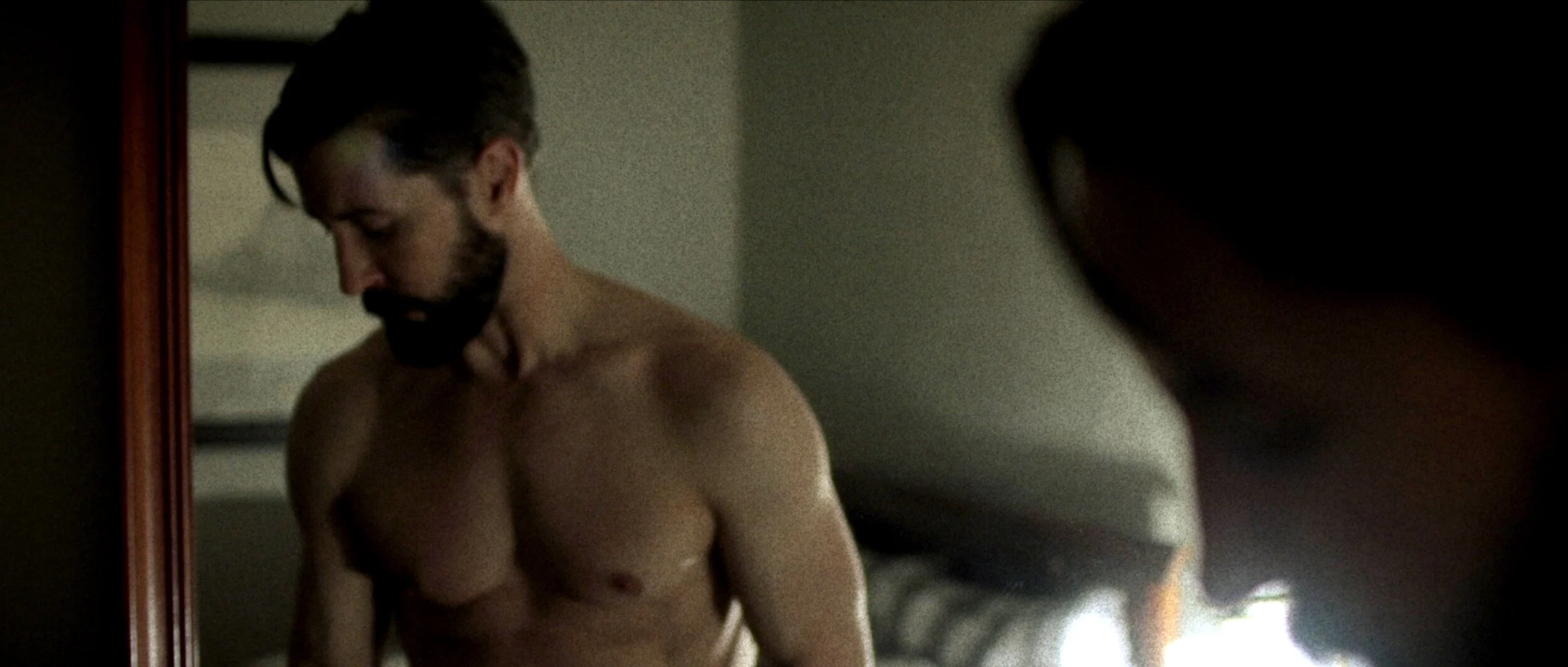 A shirtless man with a beard and glasses looking downwards, seen in a mirror in a dimly lit room.