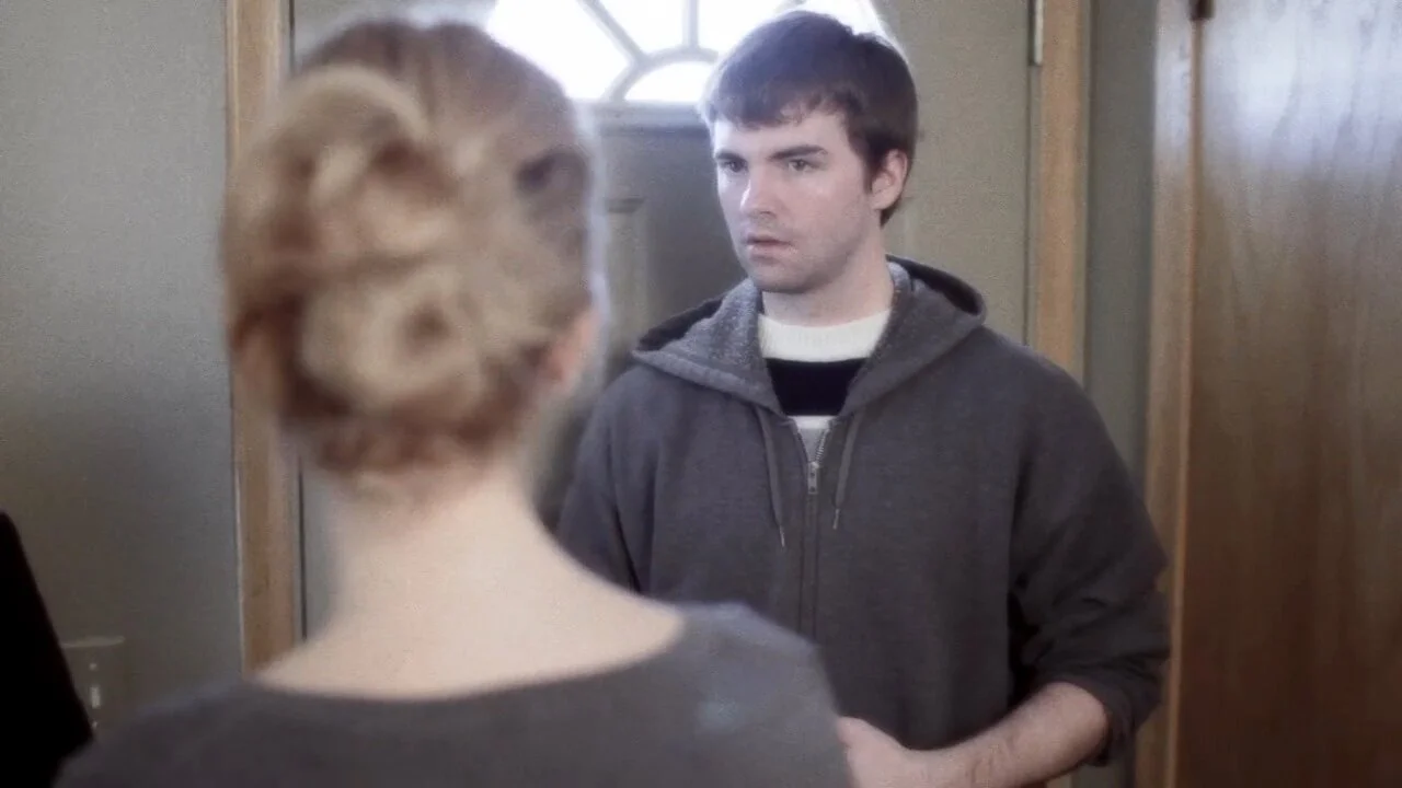 A young woman with blonde hair styled in an updo, facing a young man with dark hair, wearing a dark hoodie, as they stand in a doorway.
