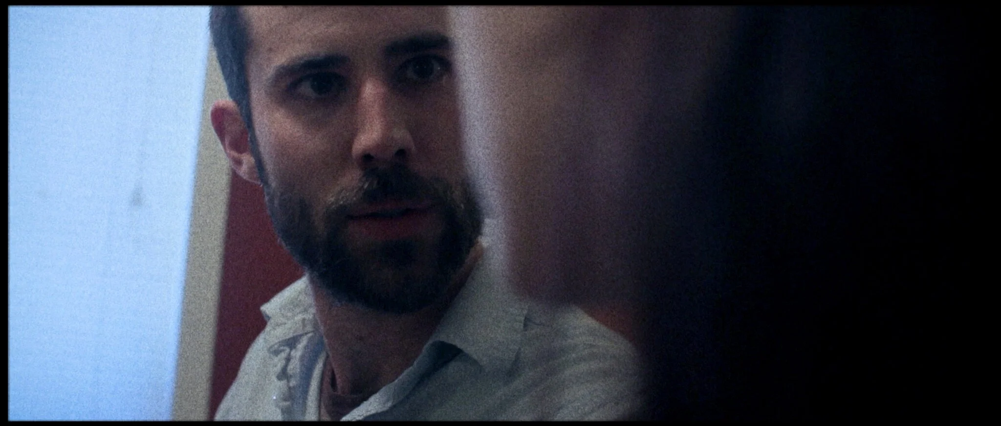 A man with dark hair, a beard, wearing a light-colored shirt, looking directly at the camera with a serious expression, in a dimly lit room with a blue wall and a red and white background.