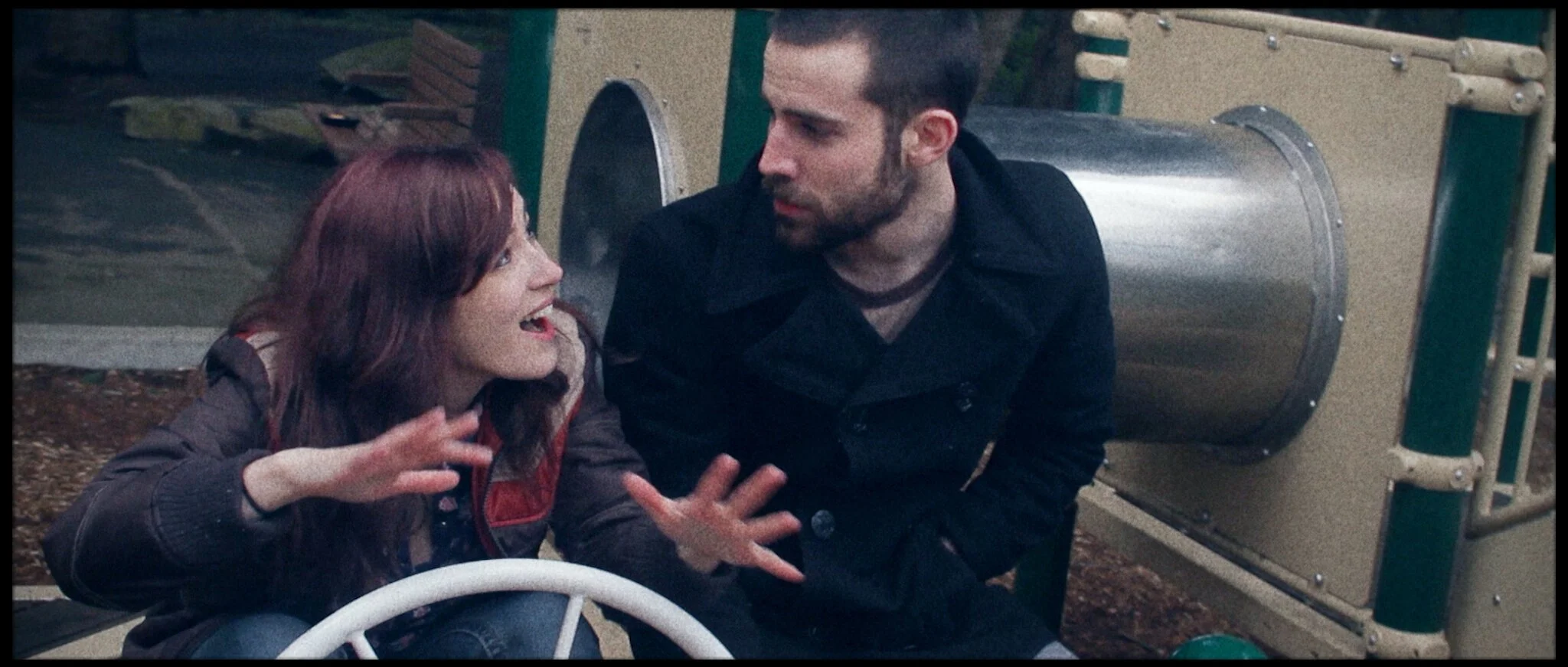 A woman with red hair and a man with dark hair and beard at a playground, both sitting on the ground near a slide, engaged in an animated conversation with expressive hand gestures.