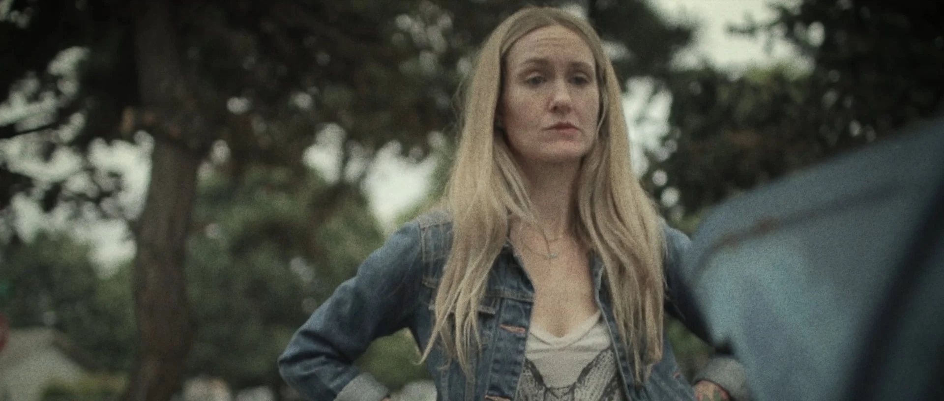A woman with long blonde hair wearing a denim jacket and a white shirt with a design, standing outdoors in front of trees, looking serious.