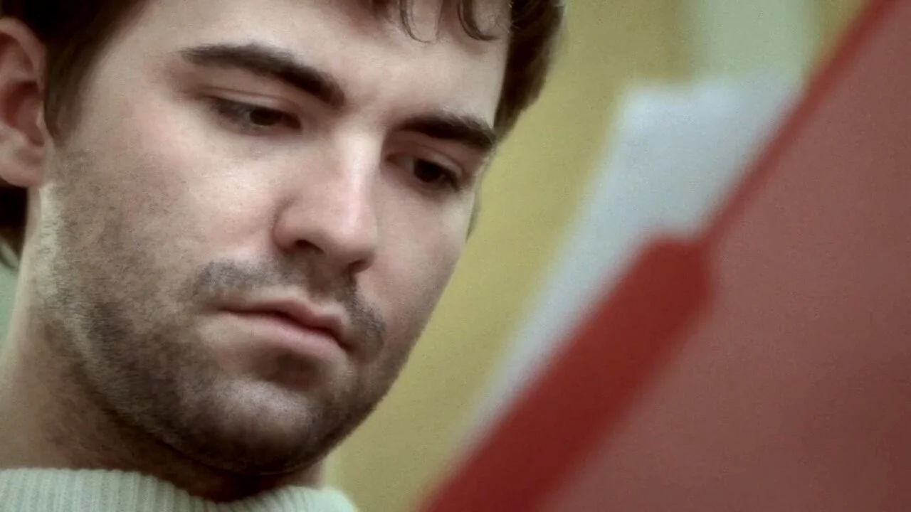 A close-up of a young man with dark hair and facial stubble looking at a red book or object in front of him.