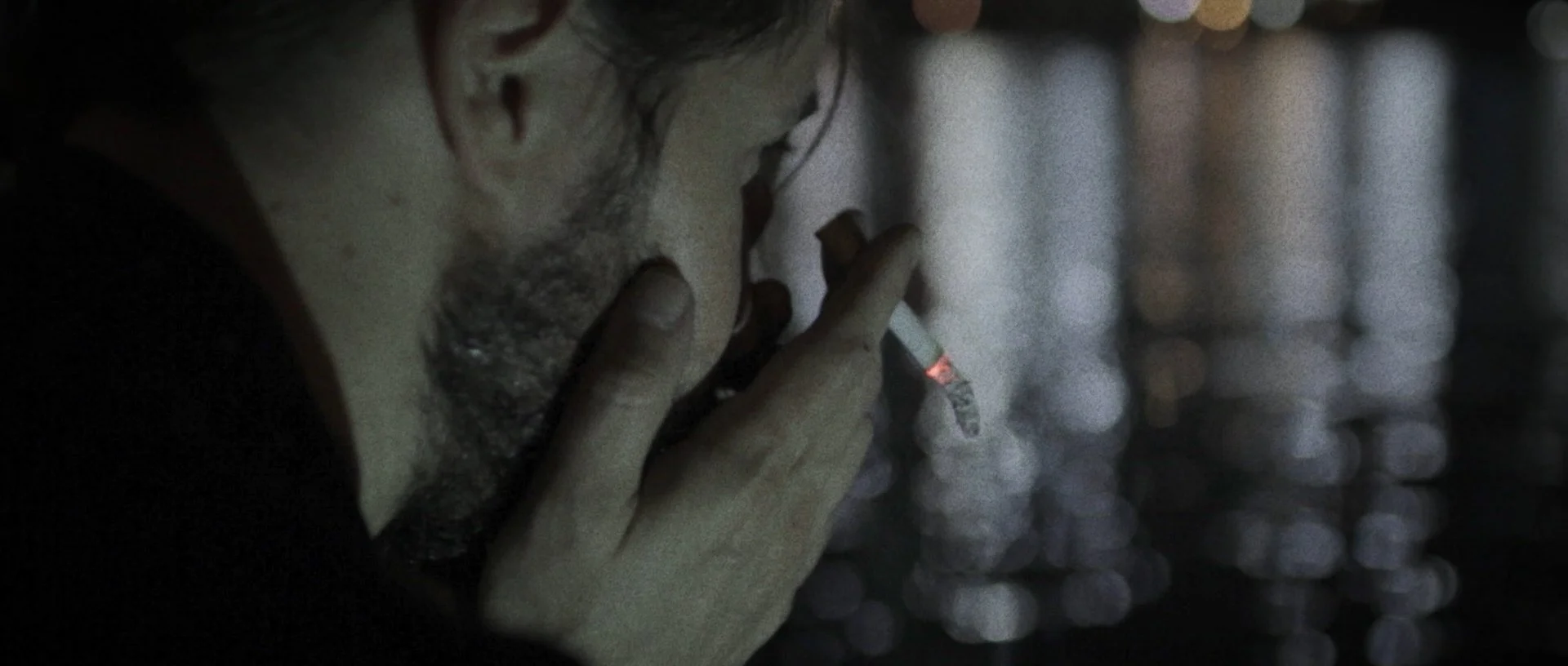 A close-up of a person with dark hair and beard smoking a cigarette in a dimly lit environment.