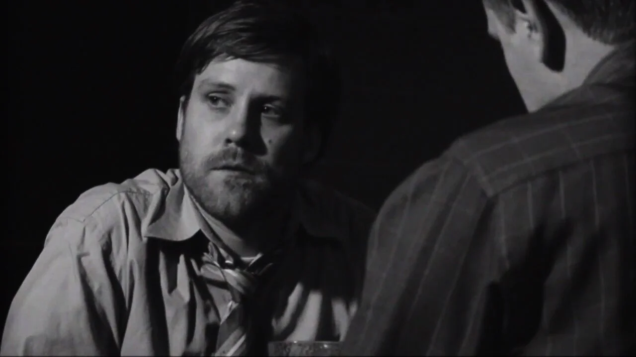 A black-and-white close-up of two men, one with a beard and thicket hair, looking serious and engaged in conversation.