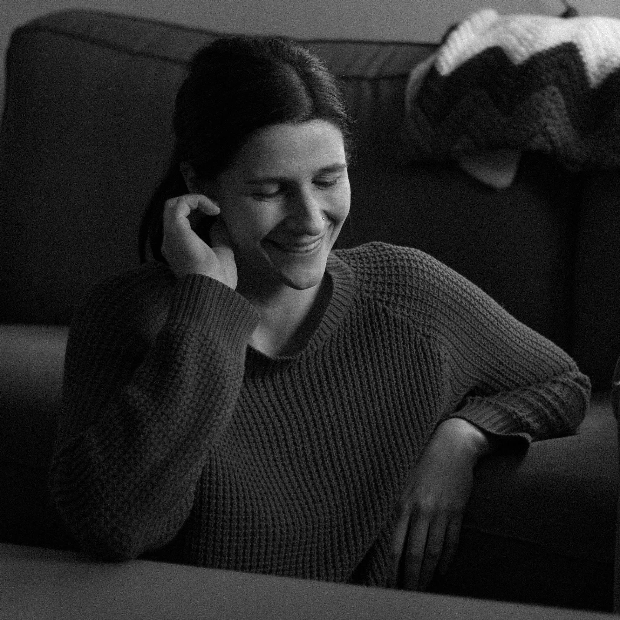 Close-up of a person's face with short hair, smiling and talking on a cell phone, wearing a knitted sweater, with a blanket folded over a couch in the background.