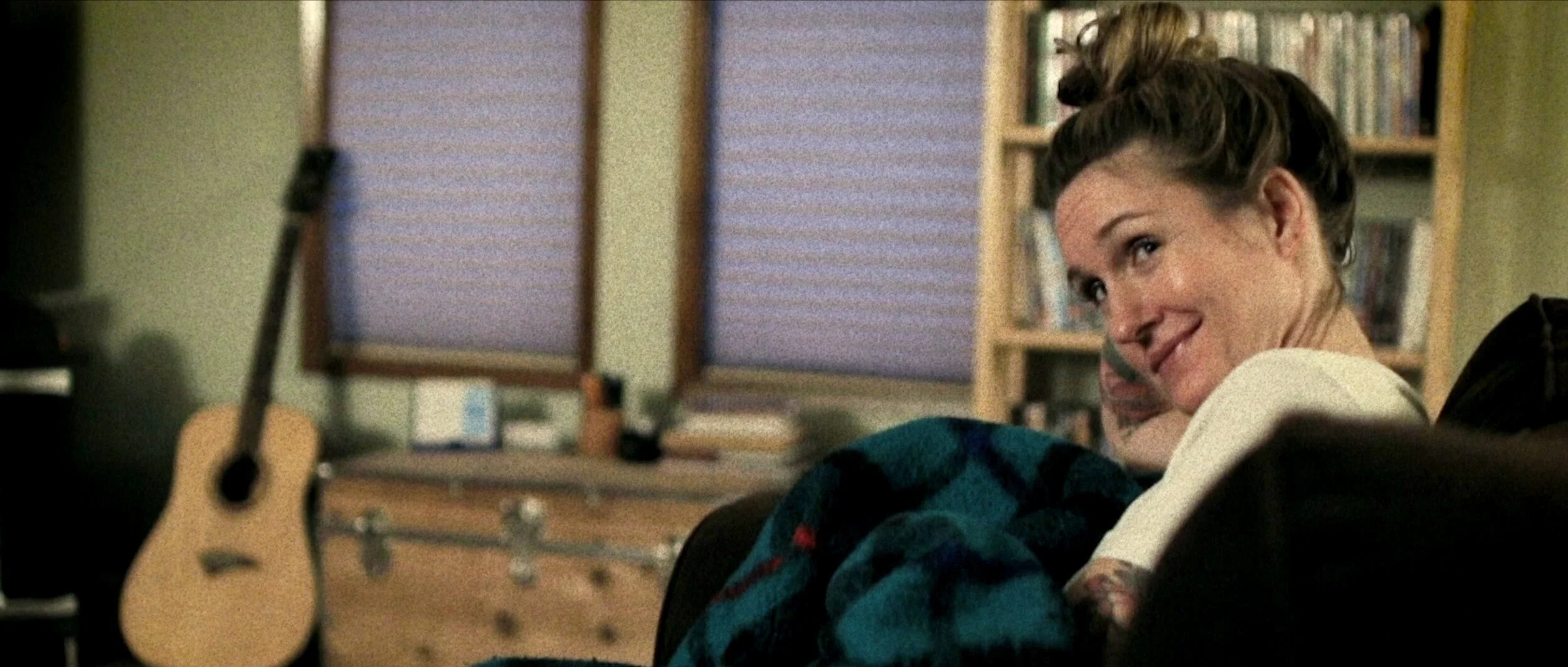 A woman with short curly hair smiling and looking over her shoulder in a cozy room with bookshelves, a window with blinds, and a guitar in the foreground.