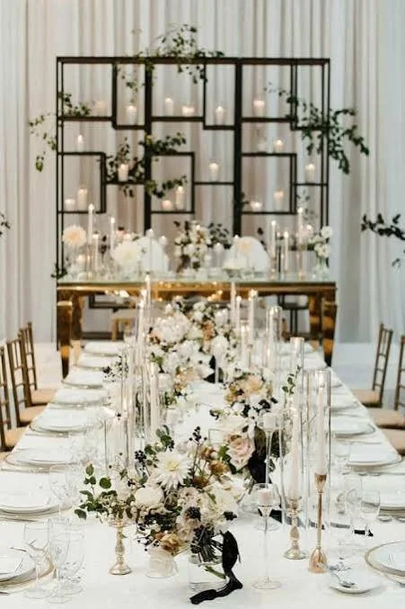 Elegant banquet table decorated with white flowers and tall candles, with a geometric black metal shelf adorned with greenery and candles in the background.