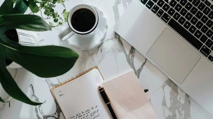 Open notebook, pen, coffee mug, laptop, and green plant on white marble surface.