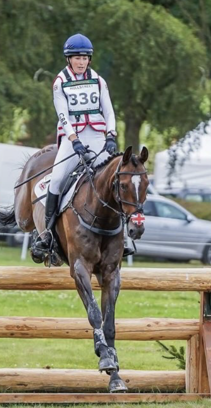 A female equestrian rider wearing a blue helmet, white riding gear with red accents, and a bib numbered 336, riding a brown horse with a white blaze and cross-shaped pattern on its nose, over wooden jumps outdoors during a competition.