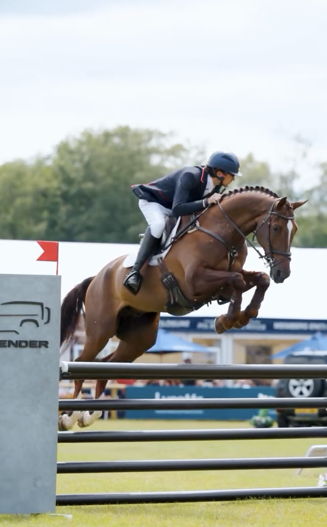 A rider in formal riding attire, including a helmet, is jumping over a horse jumping obstacle in an equestrian event.