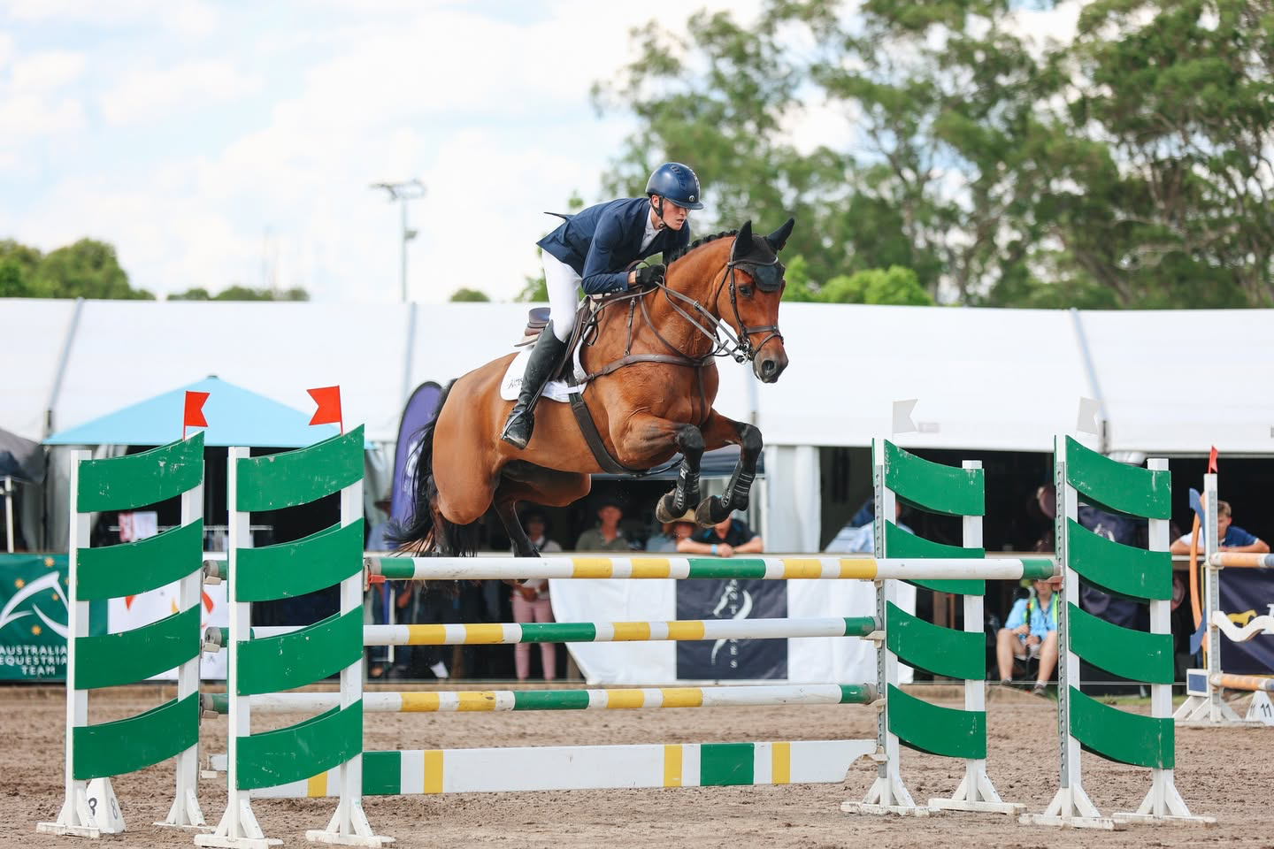 A man in a blue jacket and helmet riding a bay horse during the show jumping phase, jumping over a green and white obstacle on an outdoor  arena with tents and trees in the background.